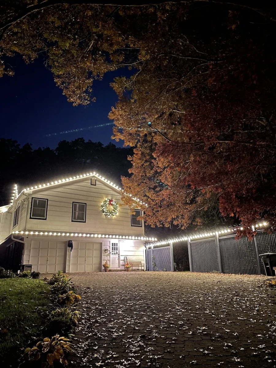 House outline and fences illuminated with C9 Chritmas lights in warm white. A big pre-lit wreath is placed in front of the house.