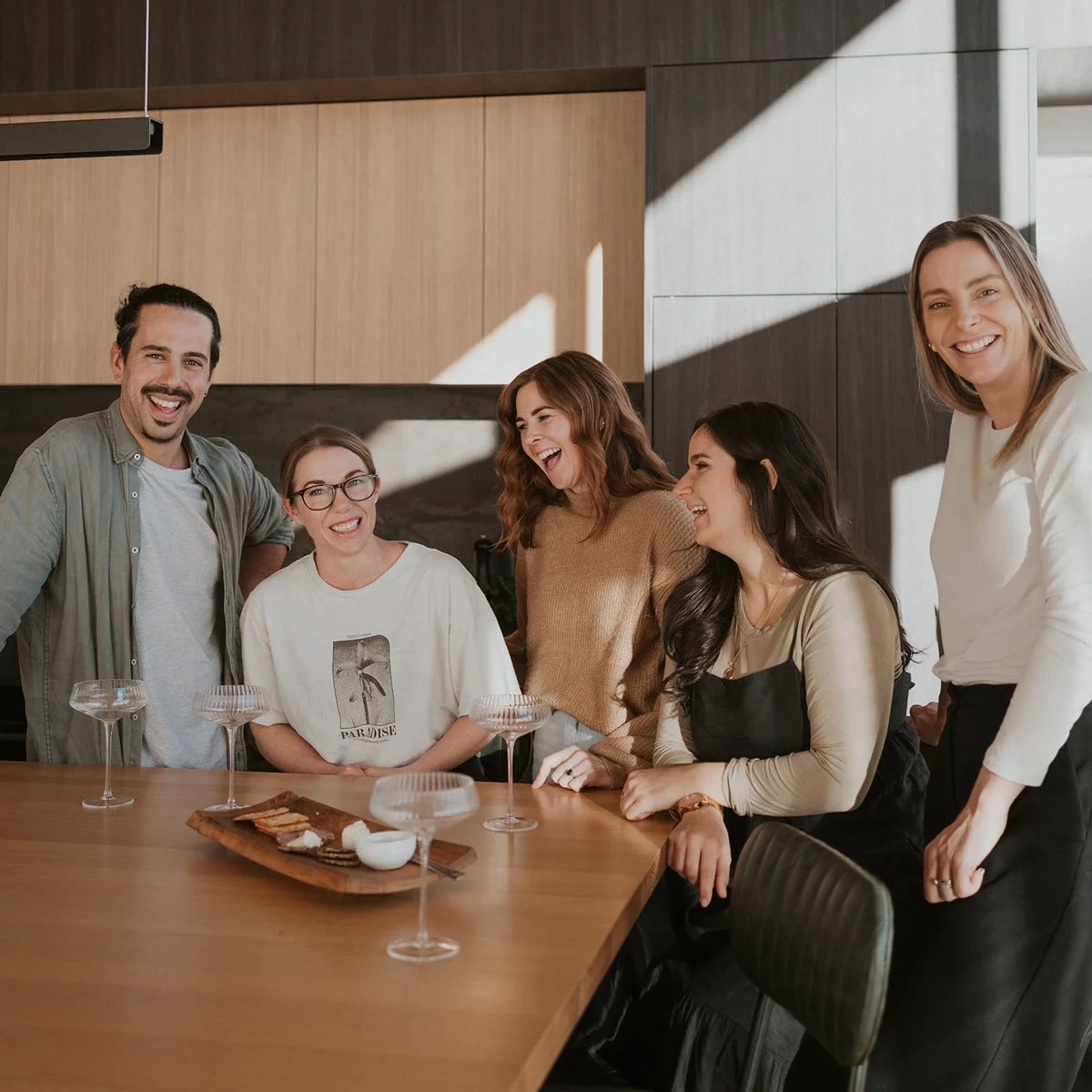 Group of five diverse friends laughing and smiling while standing around a dining table in a modern kitchen with wood cabinets and sunlight.