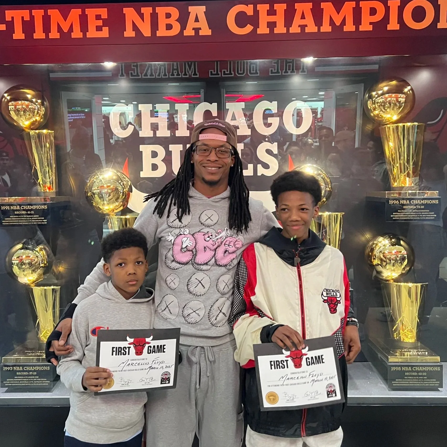 Three people, two children and an adult, standing in front of a display case with basketball trophies, holding certificates for their first NBA game, and smiling for the photo.