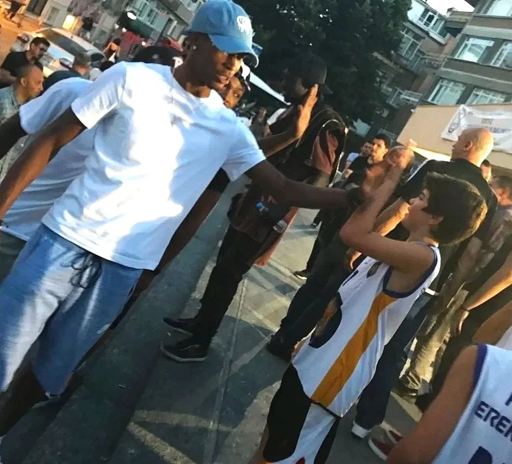 A young boy in a basketball jersey high-fives a tall man wearing a white t-shirt, light blue shorts, and a blue cap in an outdoor urban setting with several people and buildings in the background.