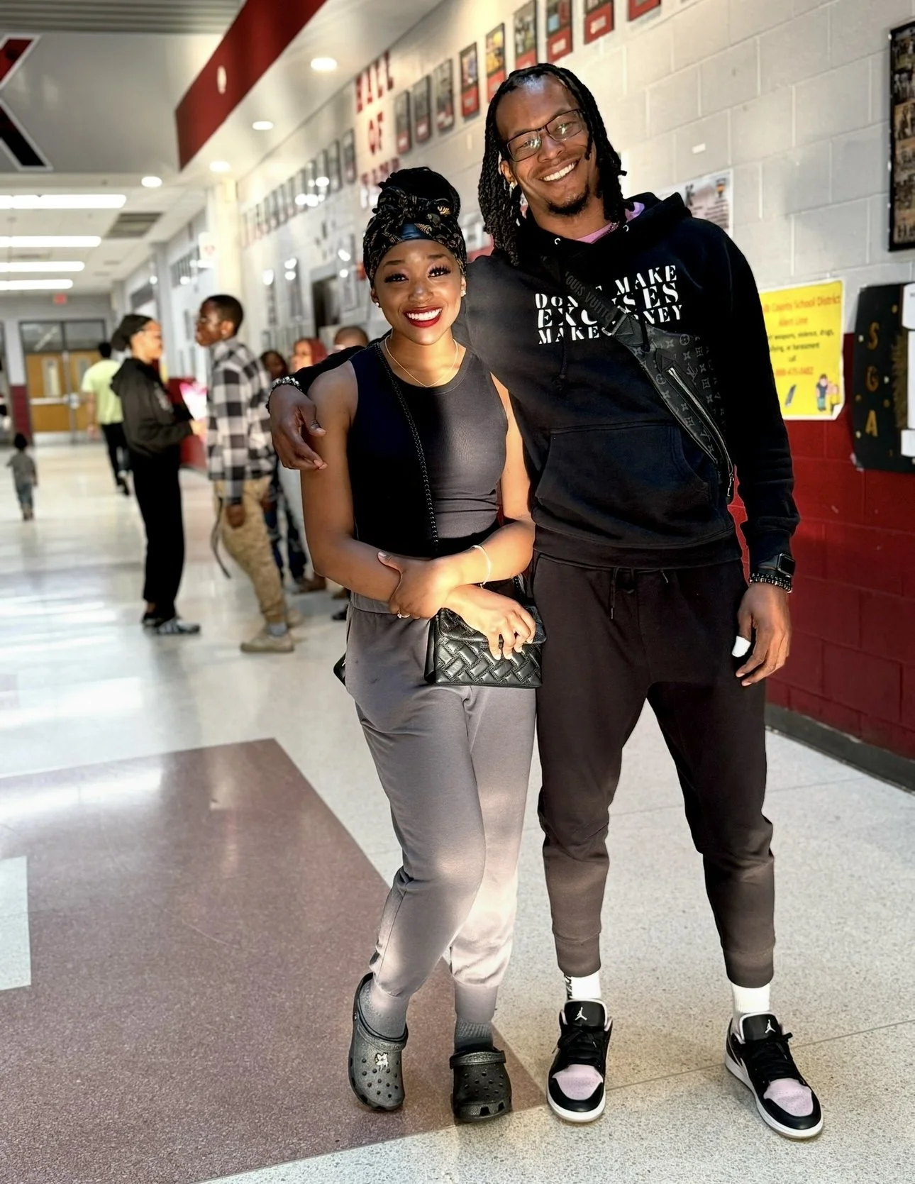 A young woman and a young man standing side by side in a school hallway, smiling and embracing. The woman is wearing a black sleeveless top, gray joggers, and Crocs, carrying a black quilted purse. The man is wearing a black hoodie, black joggers, and black and white sneakers. Several students are seen in the background, talking and walking.