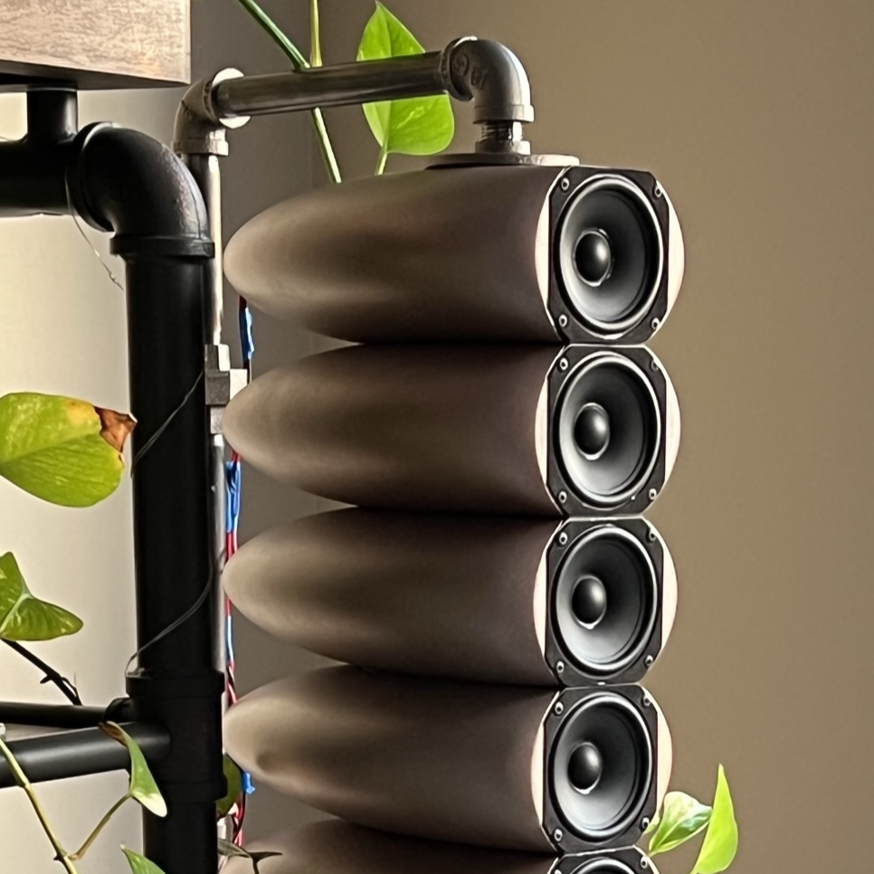 A vertical row of five large black speakers mounted on a black metal pipe structure, with green plant leaves in front of and behind the setup, against a beige wall background.