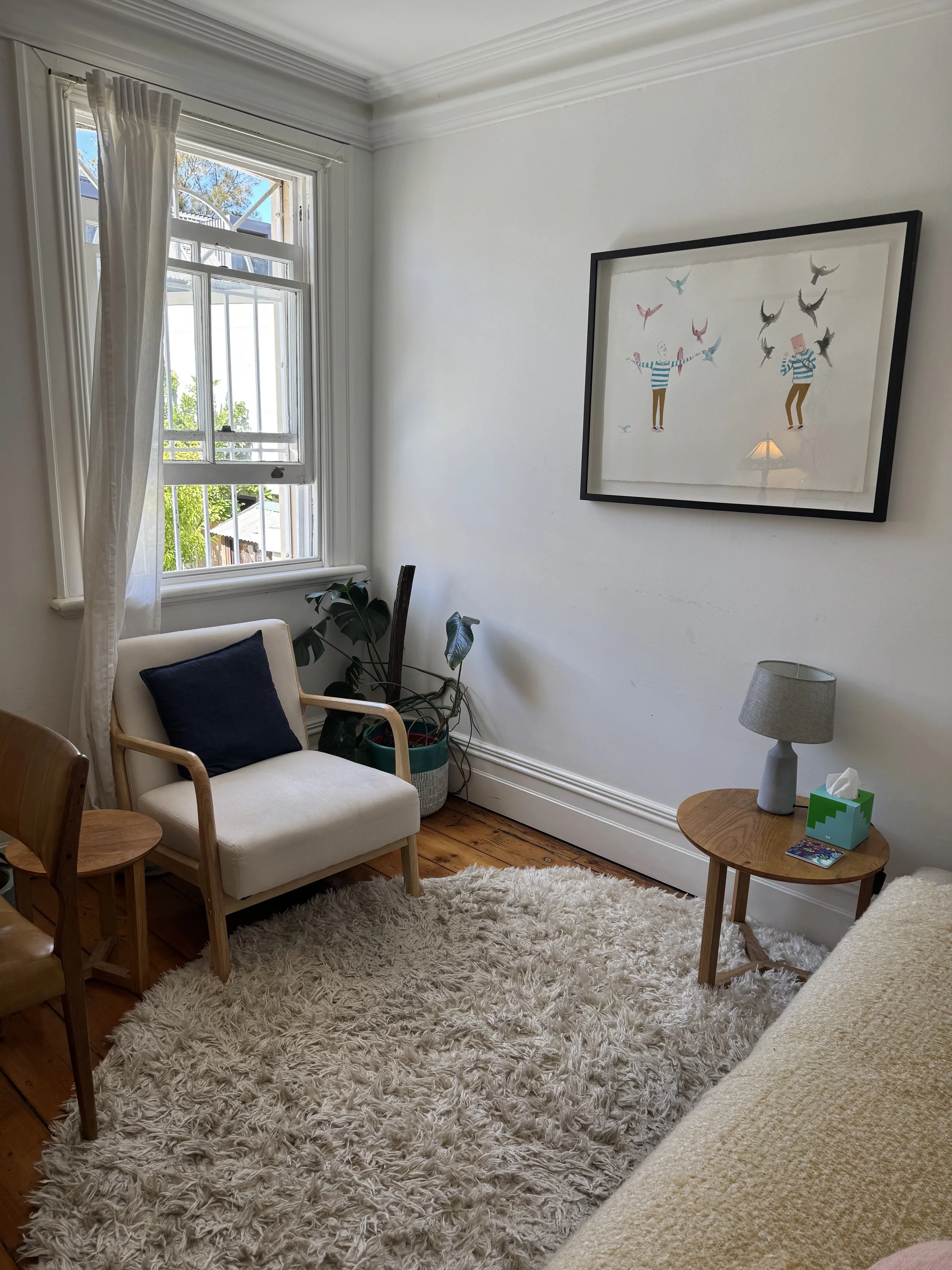 Corner of a cozy living room with a window, a white armchair with a dark pillow, a small side table with a lamp and tissue box, a fluffy cream rug, a framed whimsical artwork of two characters juggling birds, and some indoor plants.