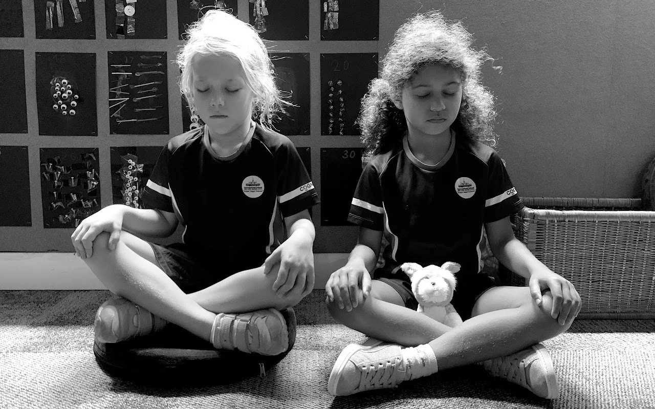 Two young children sitting cross-legged on the floor with their eyes closed, practicing meditation, one holding a stuffed animal, in a classroom.