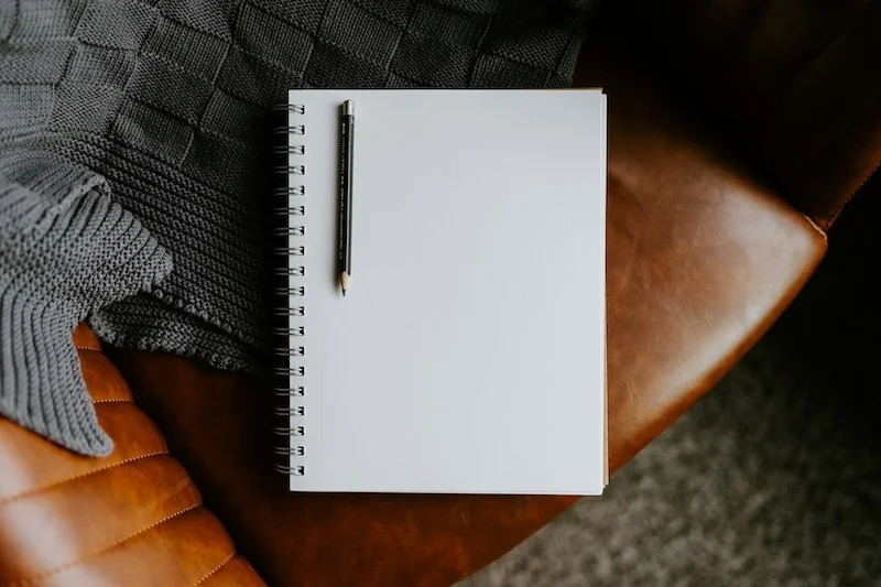 Open blank spiral notebook with a pencil resting on it, placed on a brown leather chair with a gray knitted blanket nearby.