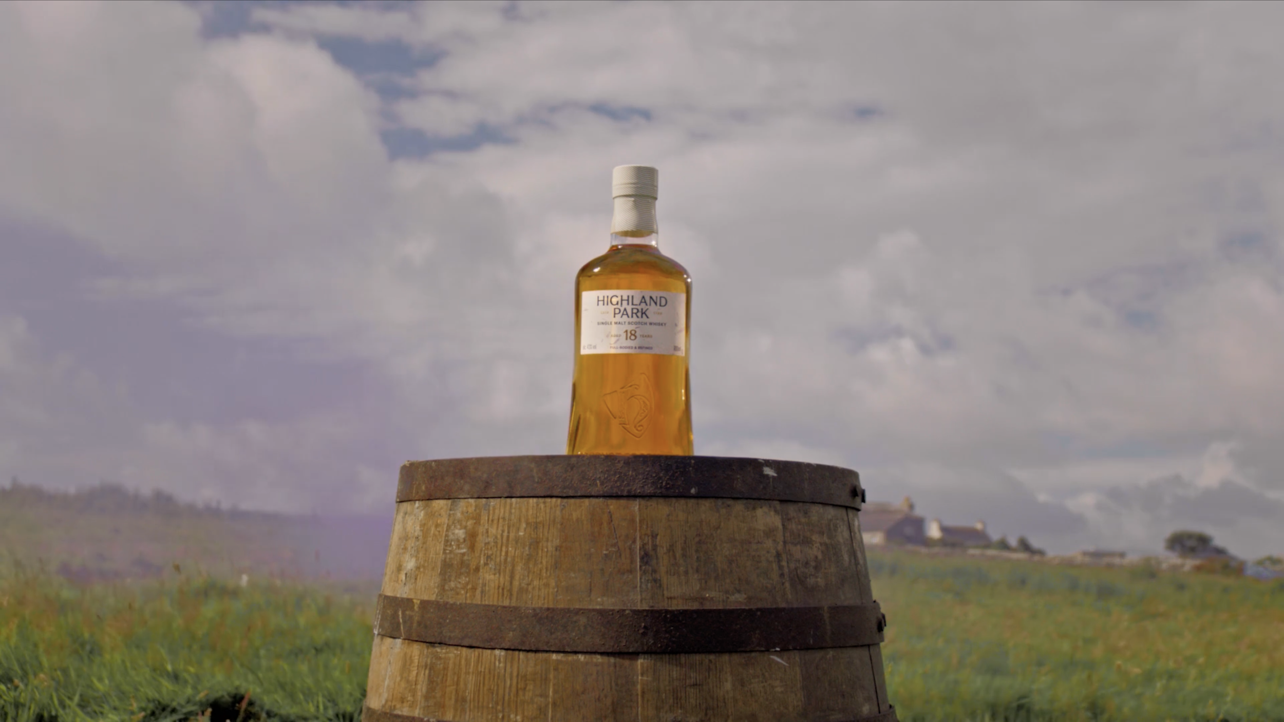 A bottle of Highland Park 18-year-old single malt Scotch whiskey placed on top of a wooden barrel outdoors, with a cloudy sky in the background.