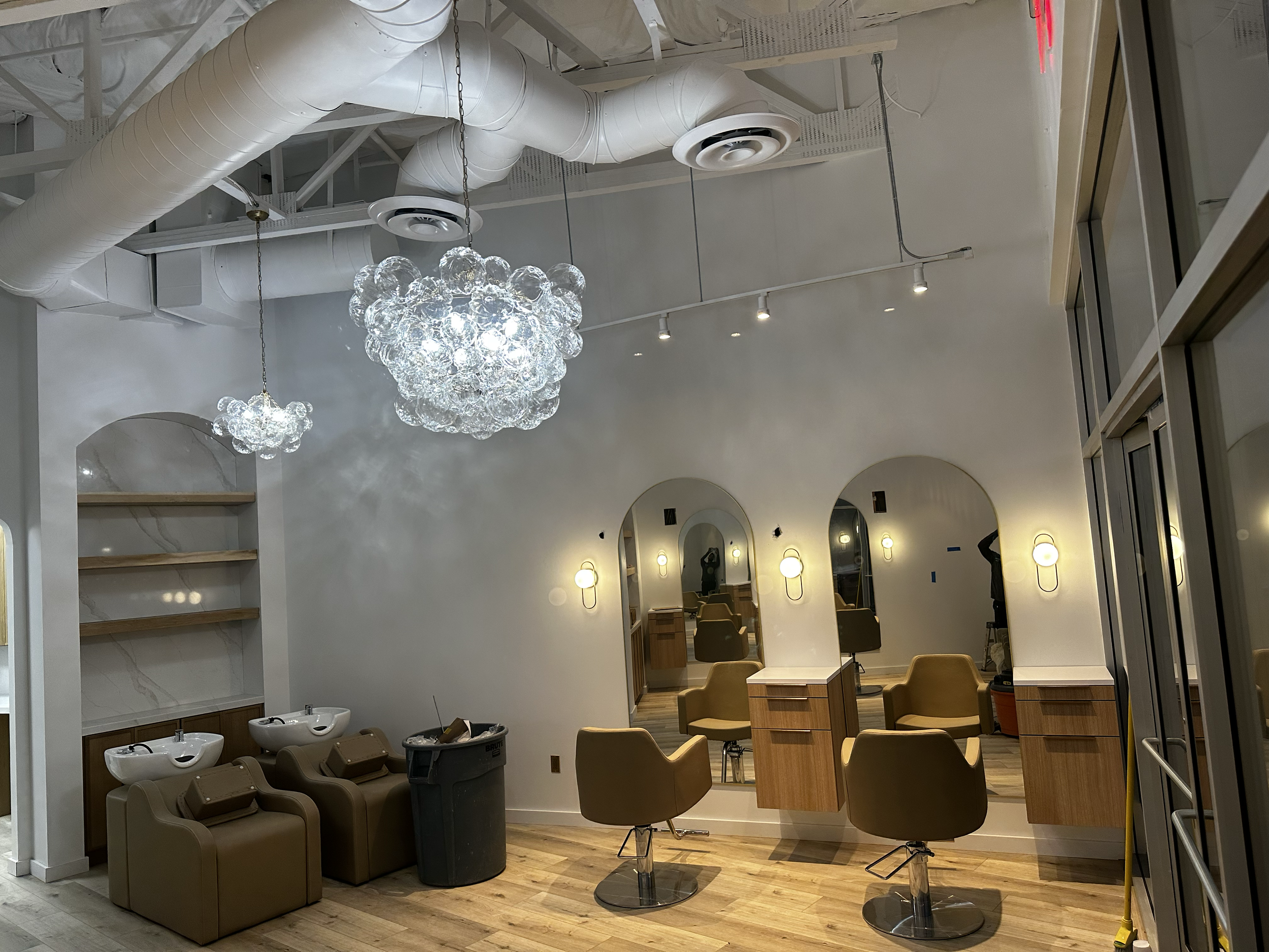 A modern hair salon with three styling chairs in front of mirrors, two used for washing hair, and a salon chair in the background. Decor includes hanging light fixtures, wooden flooring, and a neutral color scheme.