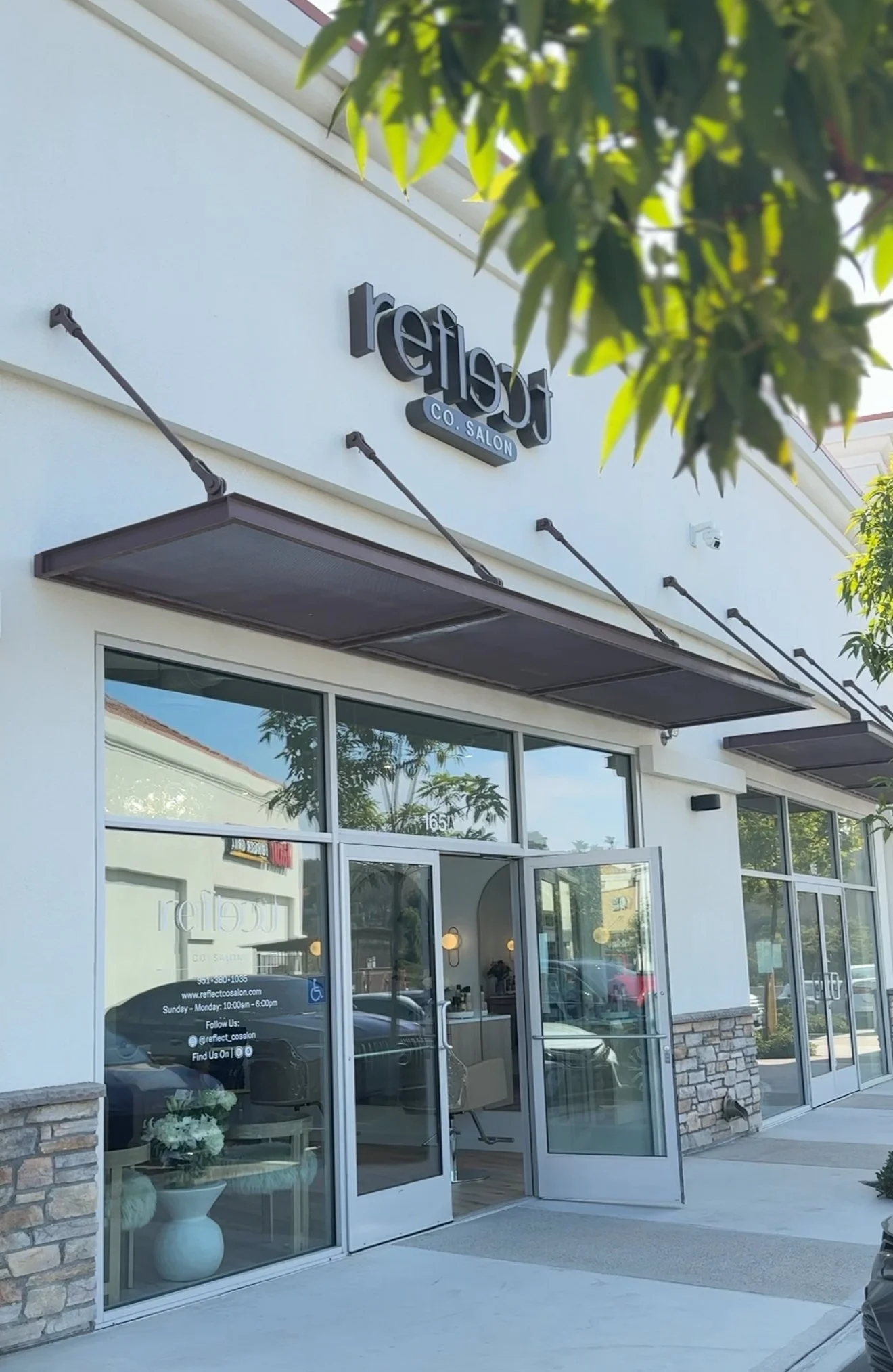Front exterior of a salon named 'Reflekt Co. Salon' with large glass windows and an open door, showing inside lighting and interior decor, with a tree partially blocking the view at the top and sidewalk in front.