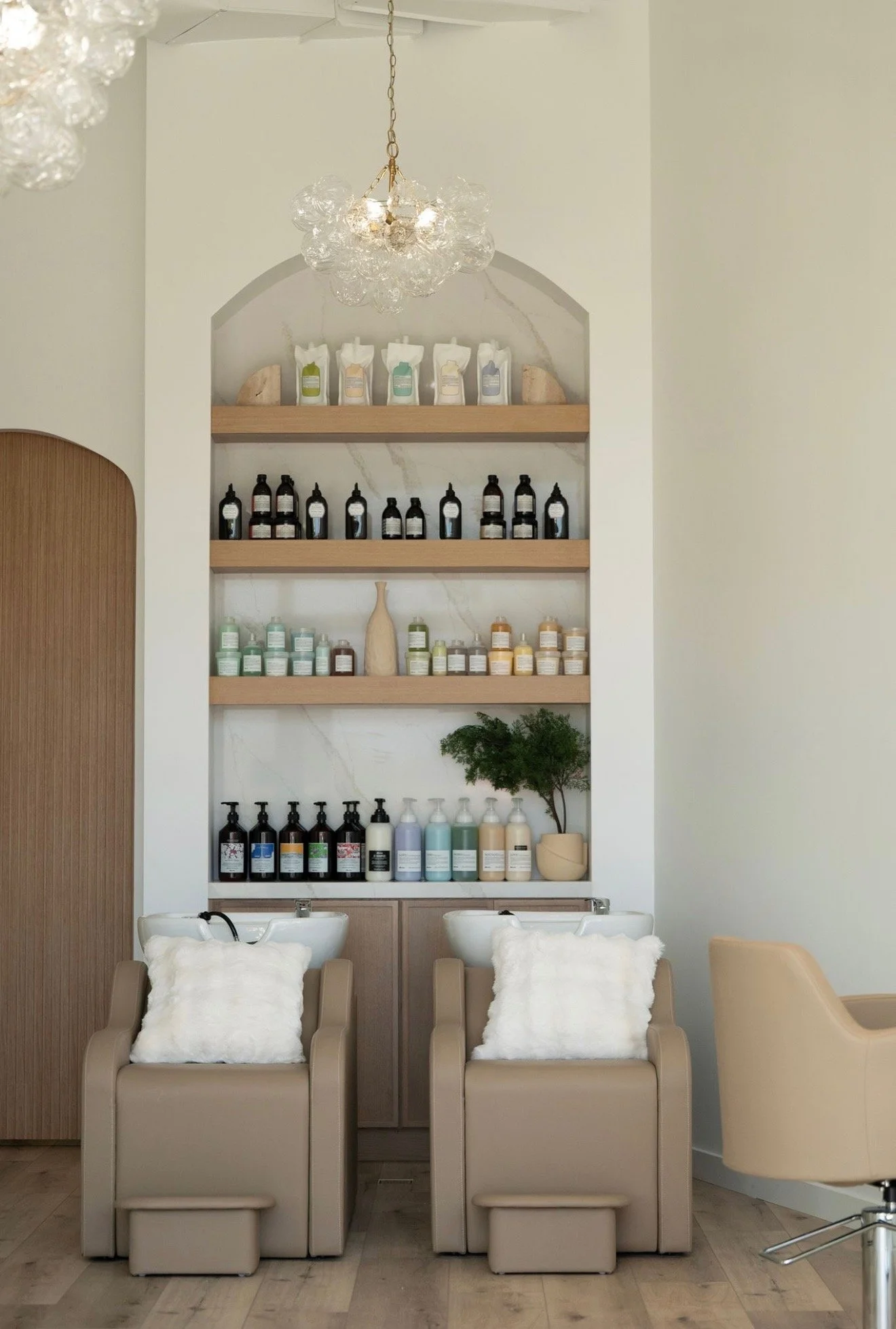 Stylish indoor salon with two beige pedicure chairs, each with white fluffy pillows, positioned in front of sinks. Behind are shelves filled with various bottles and containers of hair or skincare products, a decorative vase, and a potted tree. Elega