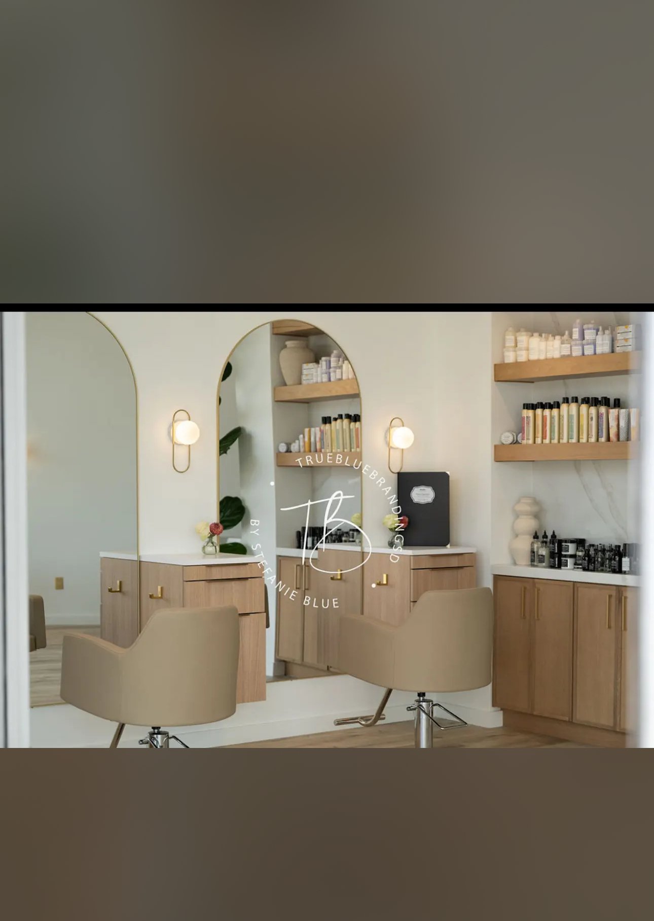 Interior of a salon or spa with beige chairs, wooden cabinets, and shelves filled with skincare or beauty products. A mirror with the words "True Blue Branding" and "by Stefanie Blue" is visible on the wall.