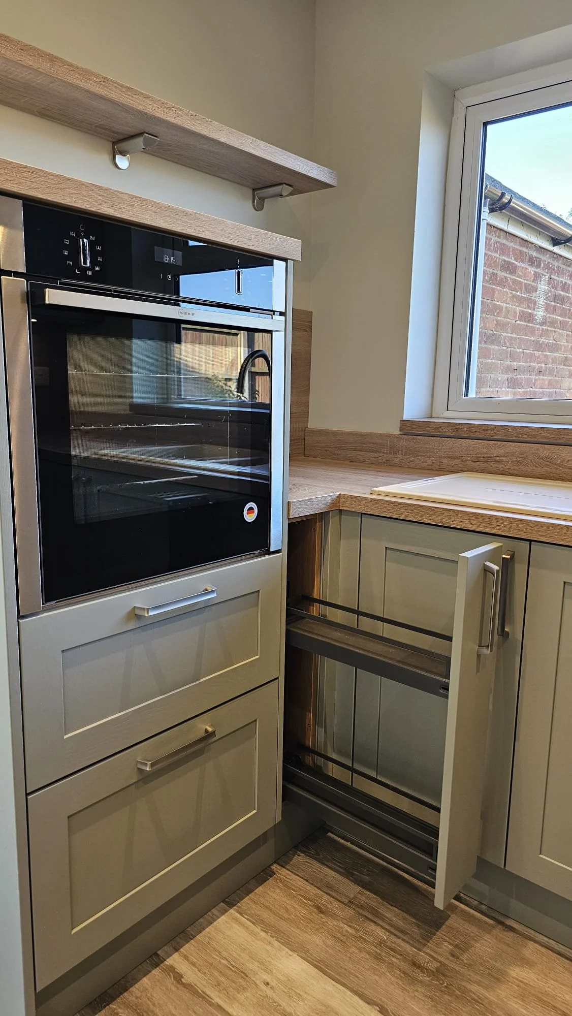 The image shows a kitchen corner with a built-in oven, gray cabinets, wood countertops, a window, and an empty pull-out spice or tray rack.