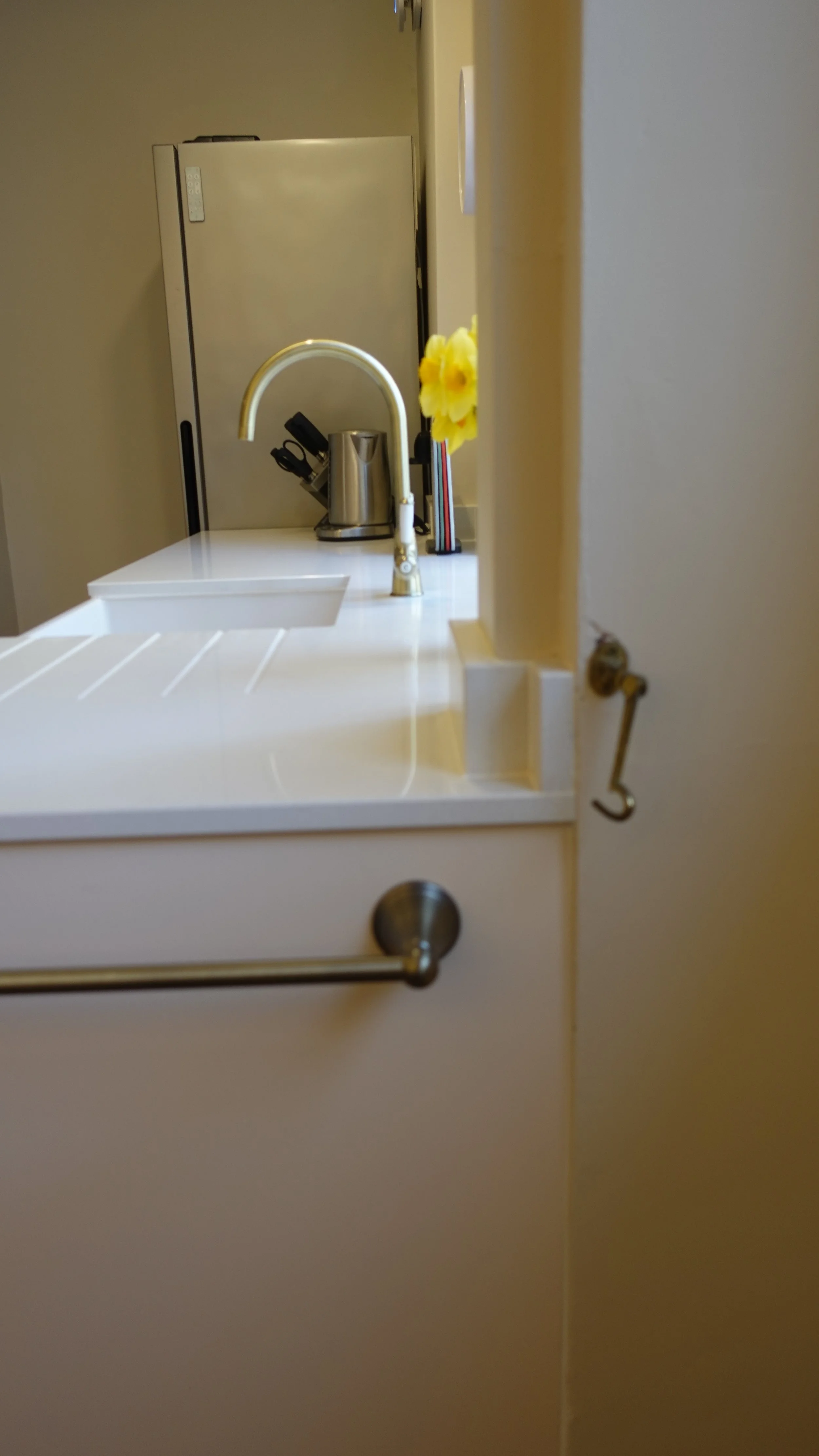 A kitchen sink with a curved faucet, a knife block, and some yellow flowers on the countertop, with a refrigerator in the background. On the side of the units a brass towel rail. 