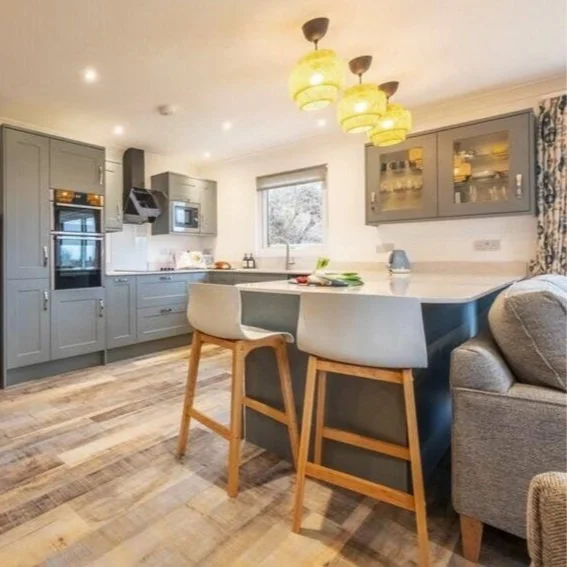 Modern kitchen with gray cabinets, a white countertop island with barstools, and yellow pendant lights. There are built-in appliances, a window, and seating area nearby.