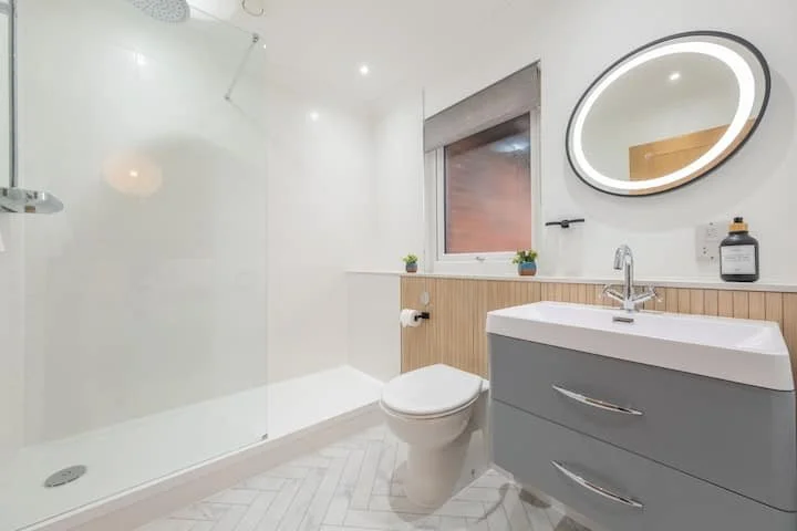 Modern bathroom with walk-in shower, toilet, gray vanity with drawers, round illuminated mirror, small window, and potted plants on the windowsill.