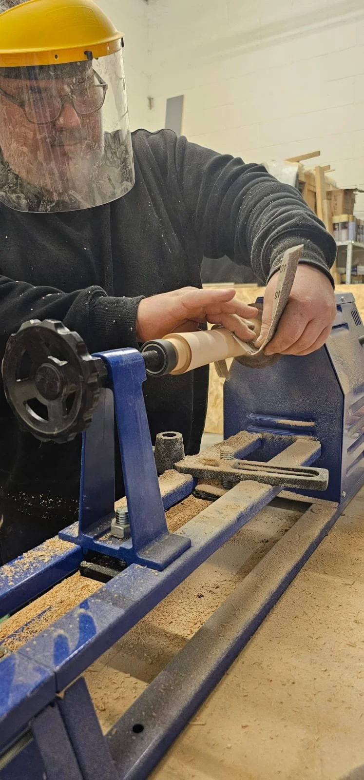 A person wearing a yellow face shield and black hoodie using a woodworking lathe machine to shape a wooden piece, in a workshop.