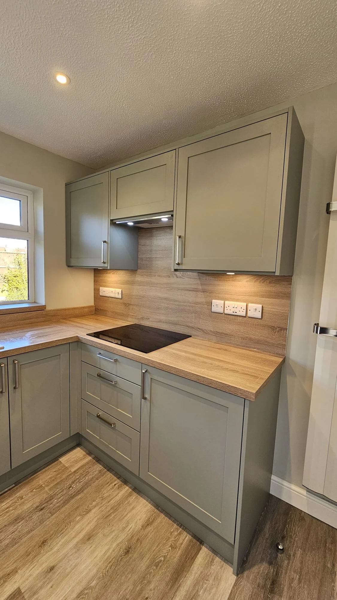 A modern kitchen with light green cabinets, wooden countertops, and a black cooktop, illuminated by under-cabinet lighting. There is a window on the left and electrical outlets on the backsplash.
