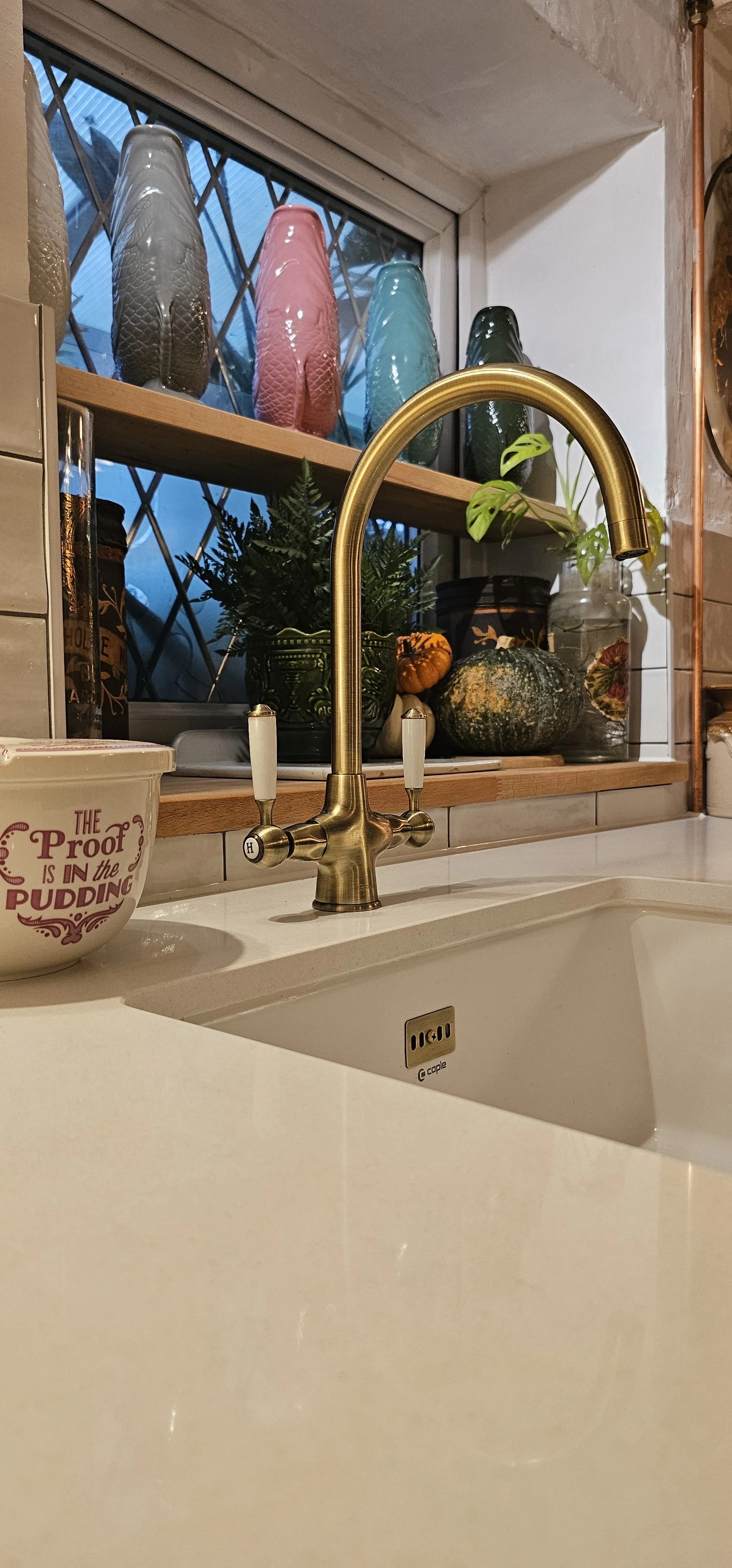 A kitchen sink with a gold-colored faucet, decorative gourds on the window sill, a basil plant, and colorful glass vases on the window shelf.