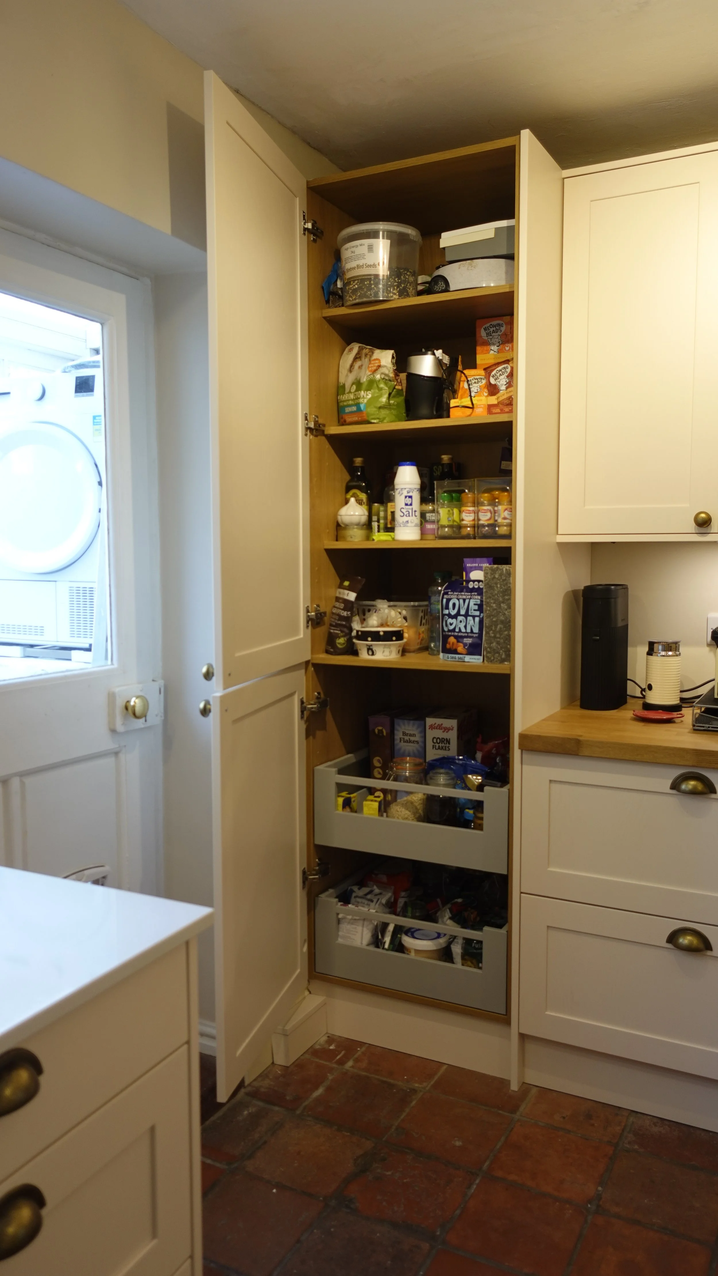 Open kitchen cabinet filled with a Space Tower storage solution by Blum various food and kitchen items, next to a countertop with small appliances, in a kitchen with tile flooring.