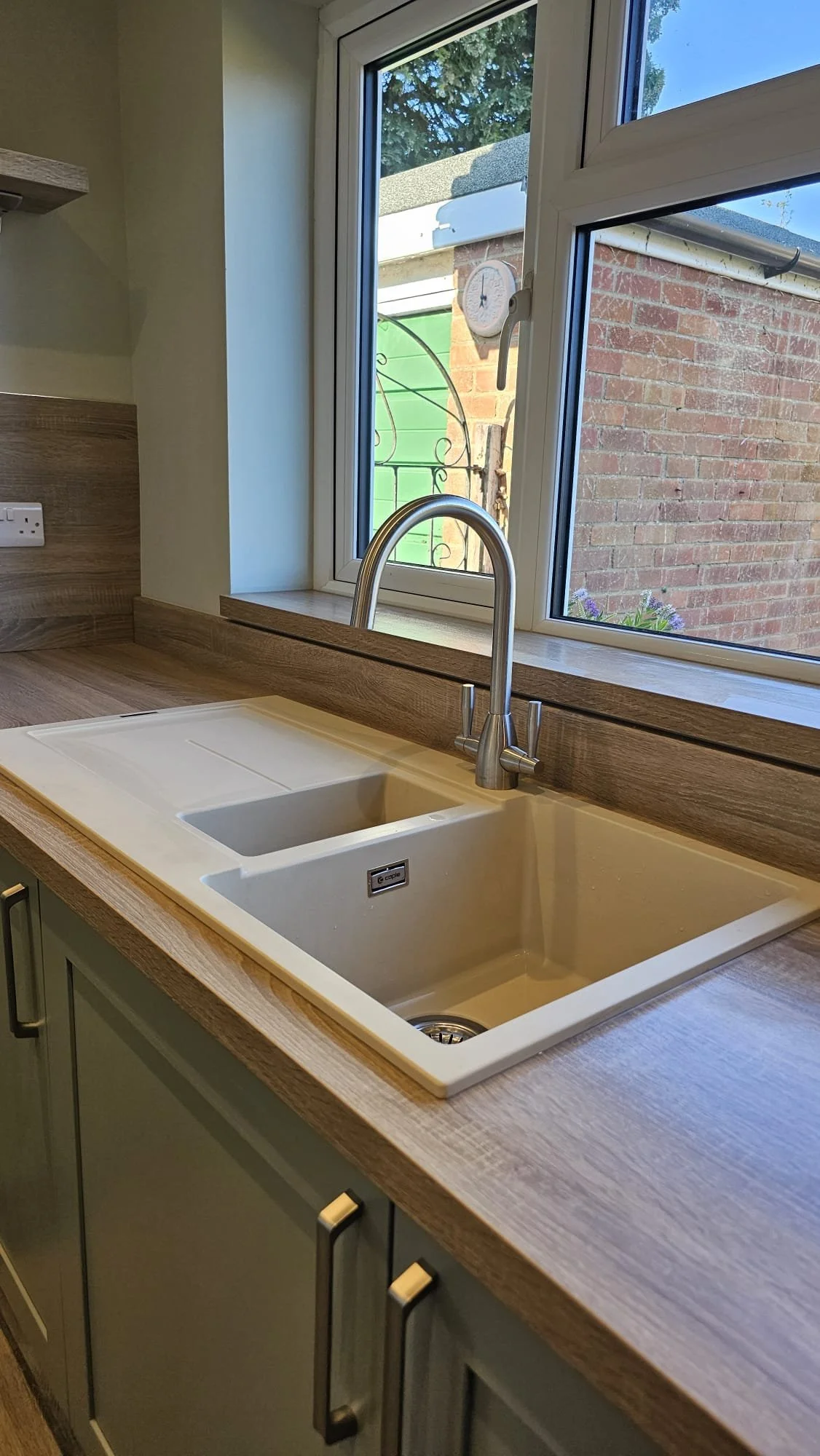 Kitchen sink under a window, with a brick house and garden visible outside.