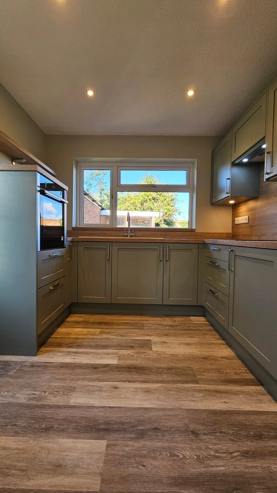A modern kitchen with green cabinets, wooden countertops, a window above the sink, and wood-look flooring.