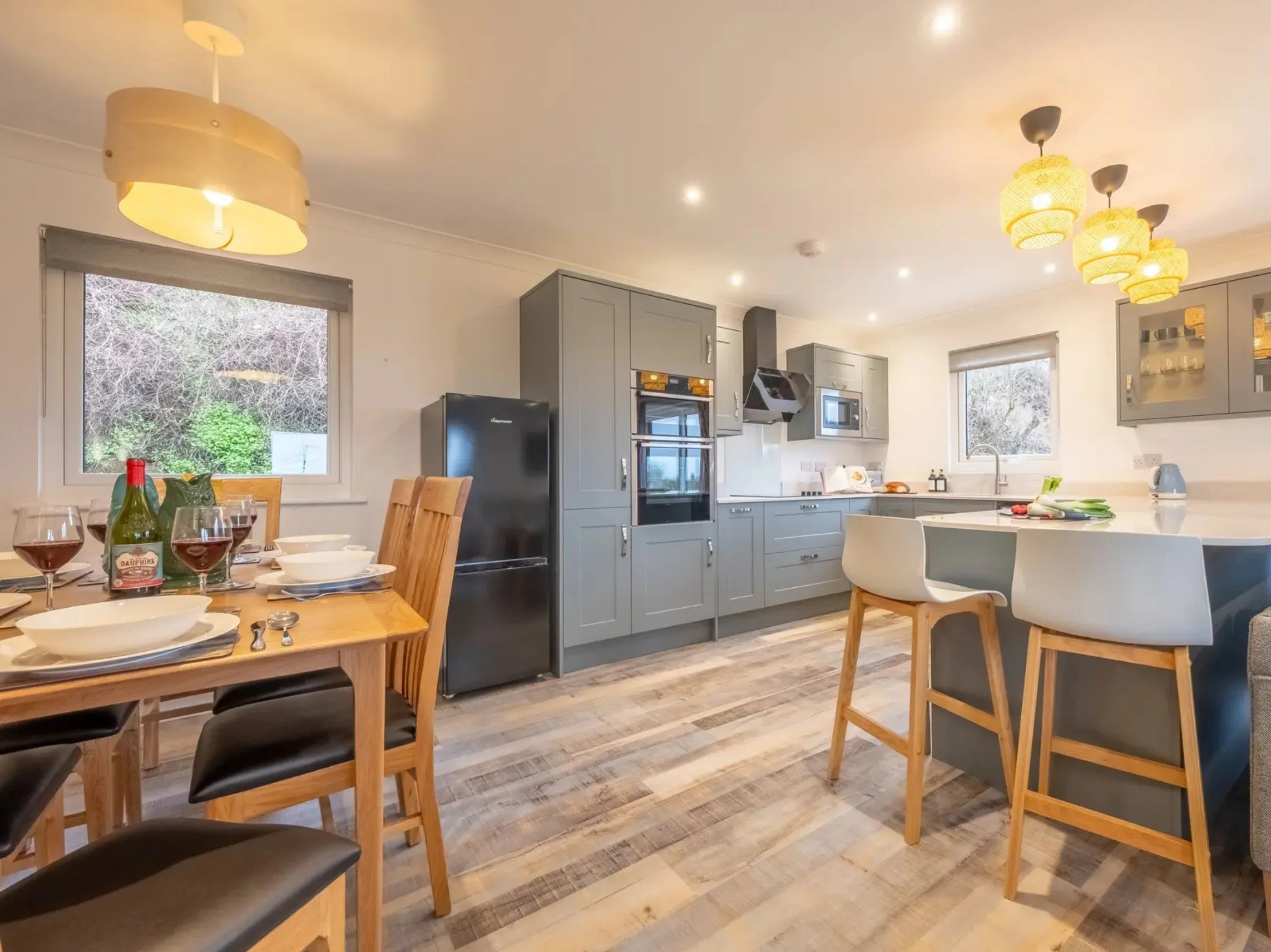 Modern kitchen and dining area with grey cabinets, wooden dining table with place settings and wine glasses, light wood chairs, white countertops, bar stools, and large windows letting in natural light.