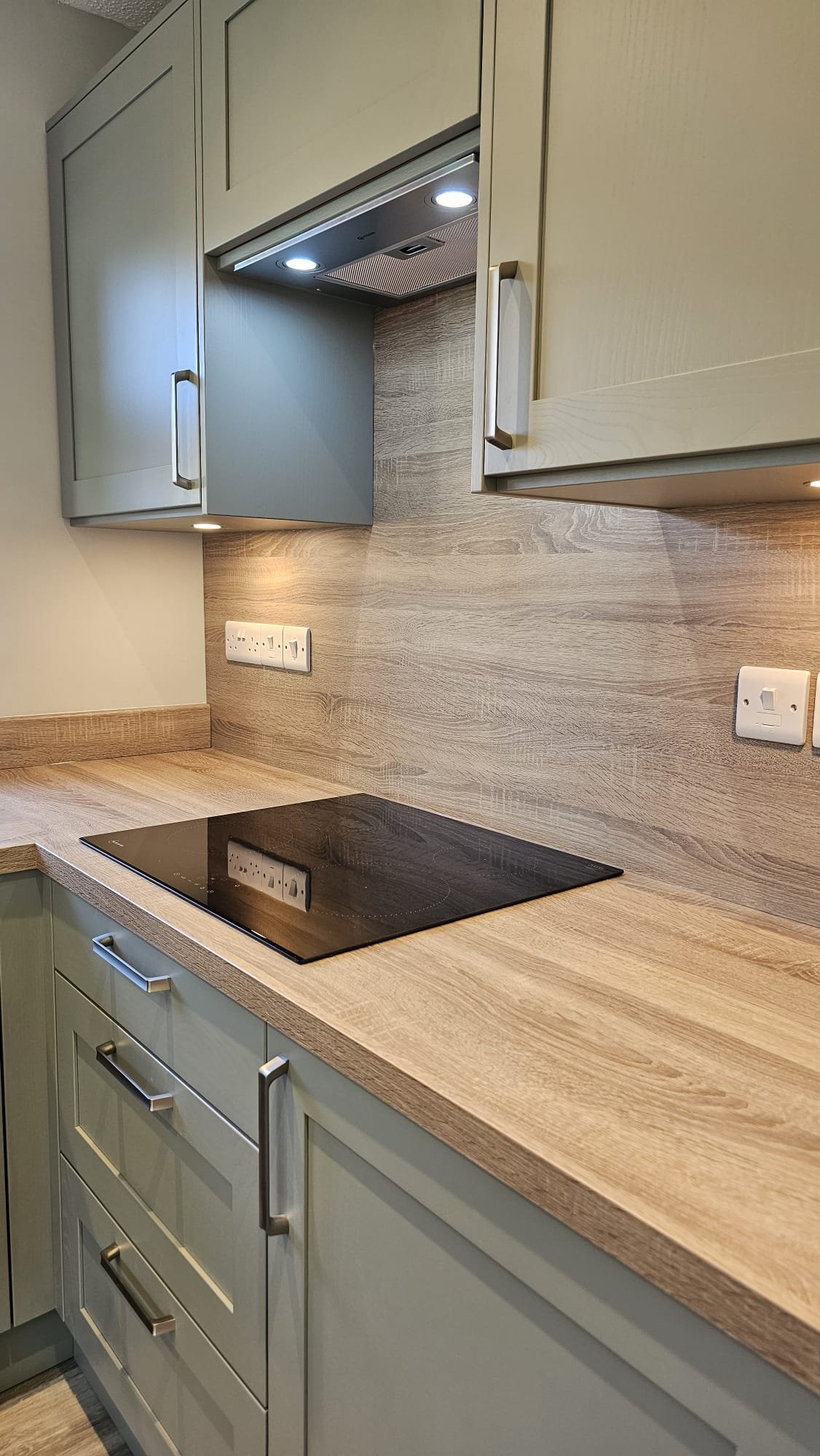 A modern kitchen countertop with a built-in electric stove, beige cabinets, a wooden backsplash, and electrical outlets.