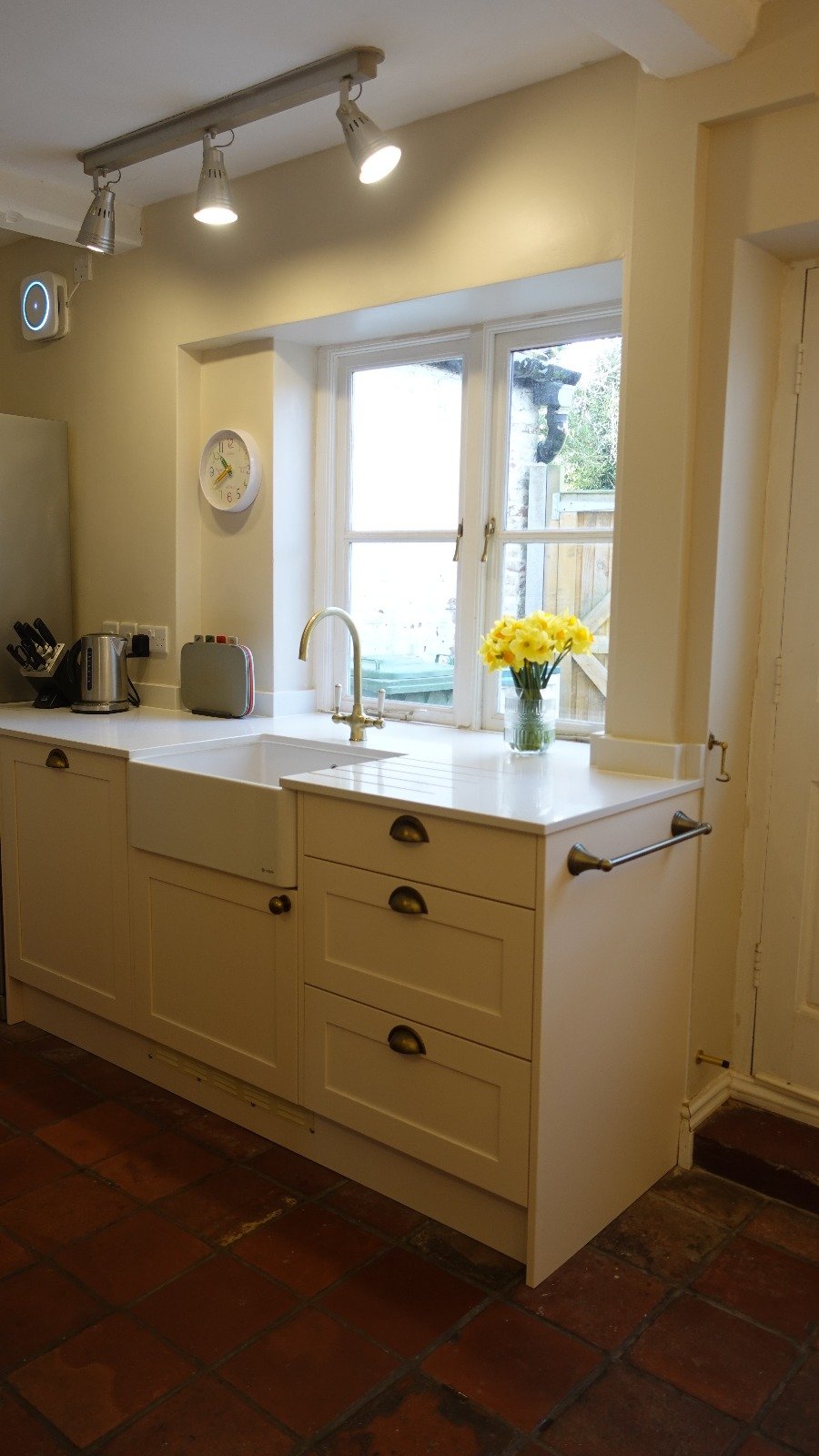 Kitchen with white cabinets, a single basin farmhouse sink, a window with a vase of yellow flowers, wooden floor tiles, and modern ceiling lights.