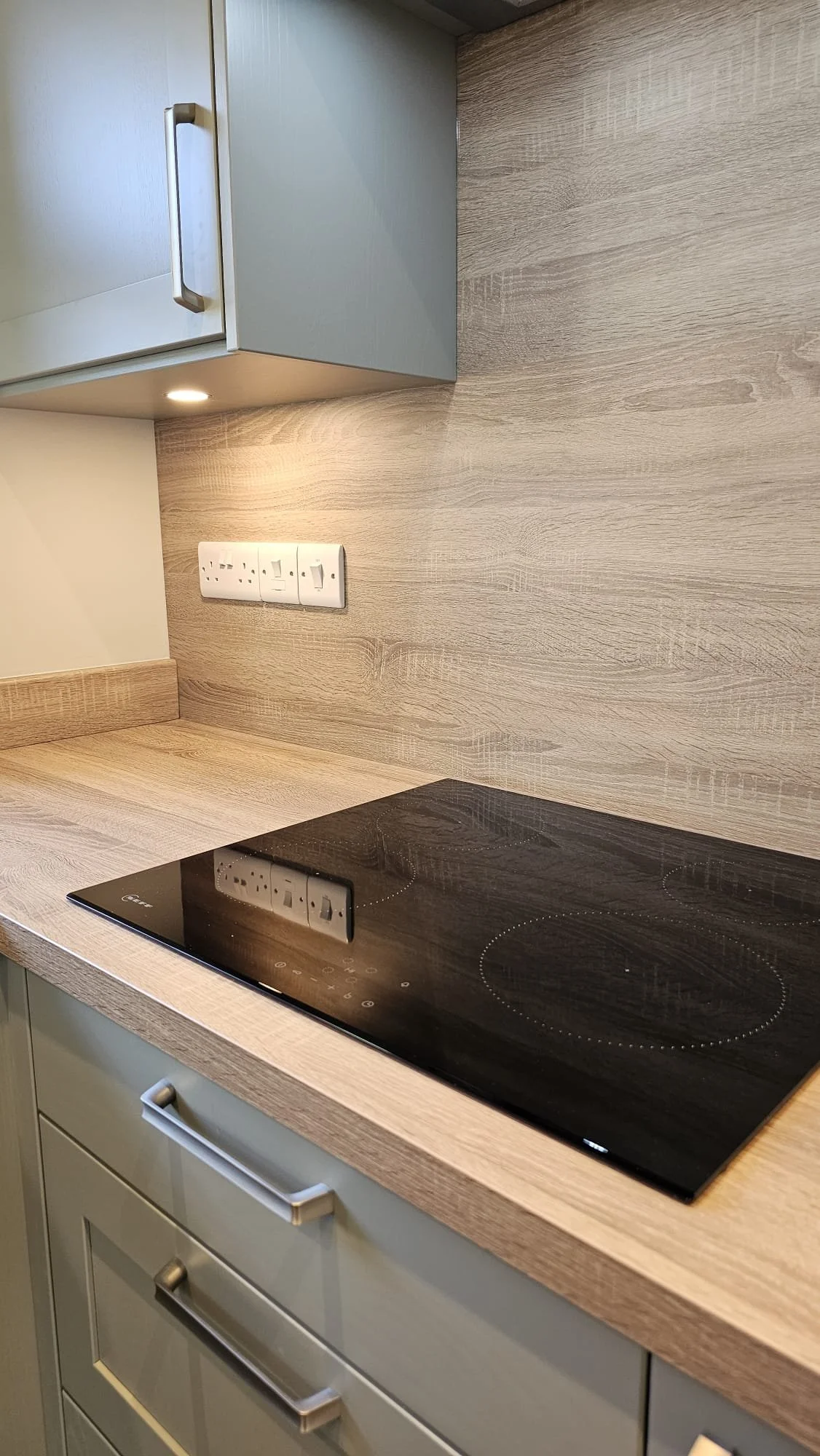 Kitchen countertop with built-in black electric stove, electrical outlets, and a light-colored wooden backsplash and cabinetry.