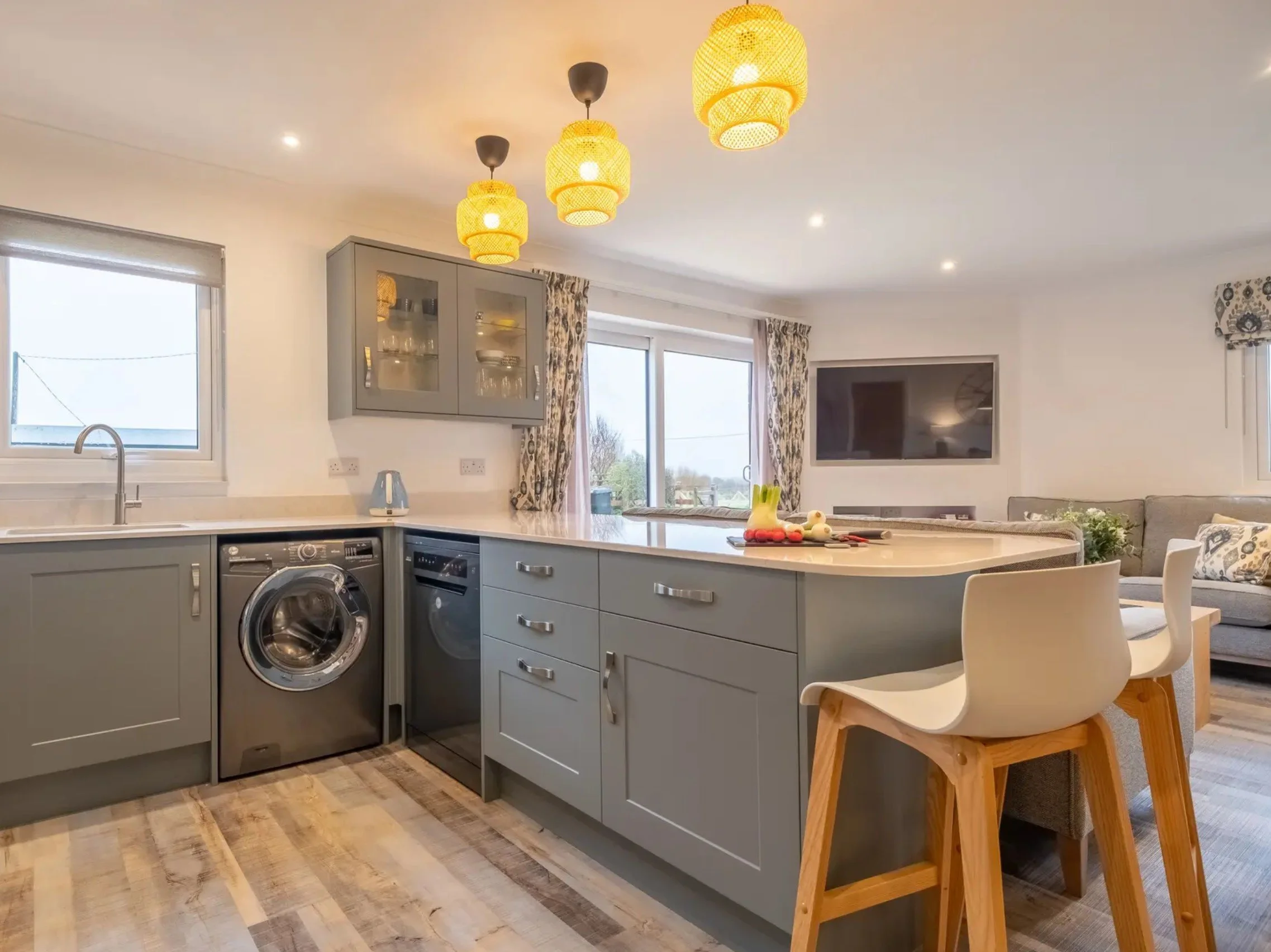 Bright, modern kitchen with gray cabinets, a white countertop, and laundry appliances. Hanging yellow pendant lights and floral curtains enhance the space, with a view of the living room in the background.
