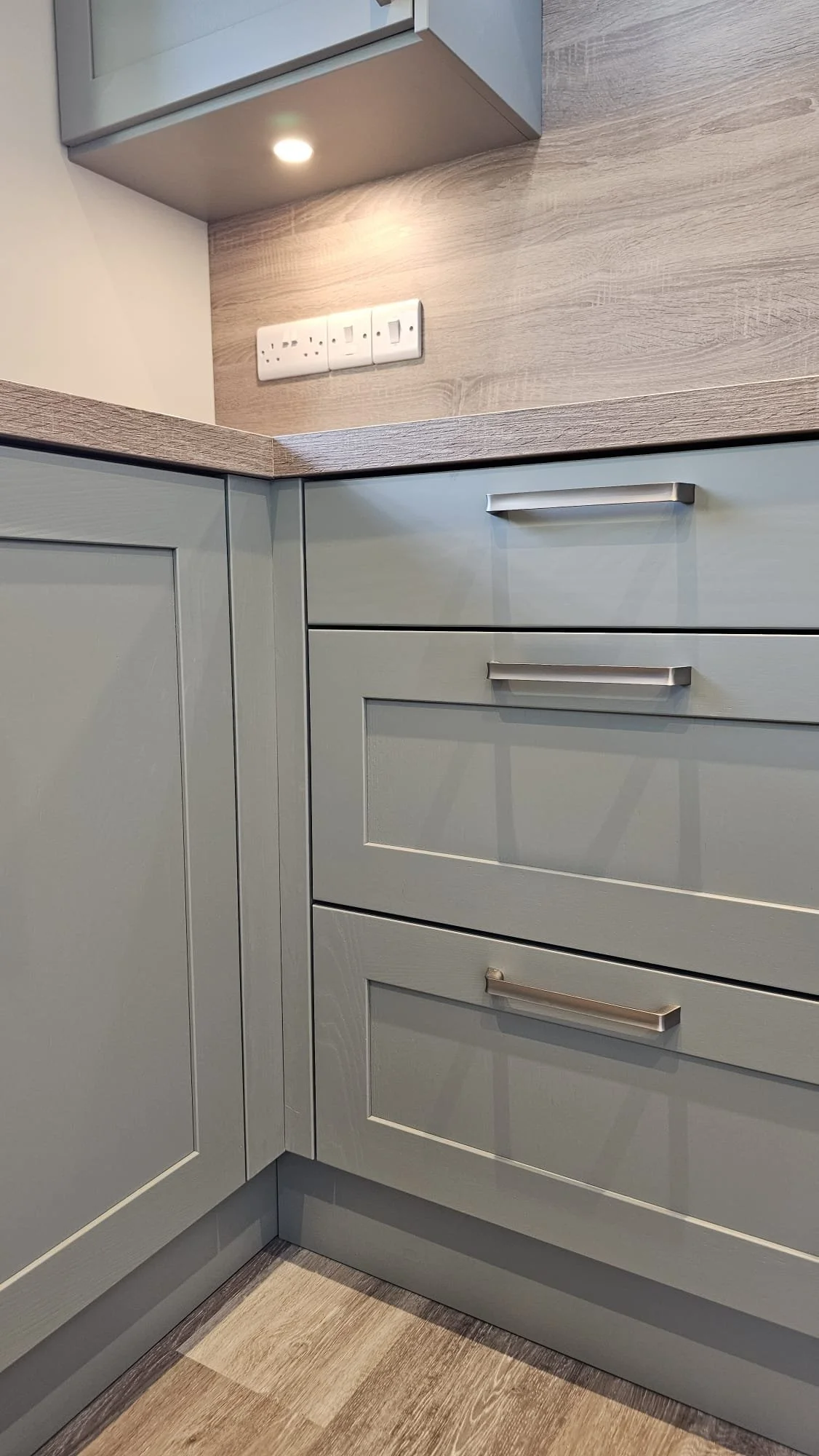 Close-up of light green kitchen cabinets with silver handles, wooden countertop, and a wall with a power outlet.