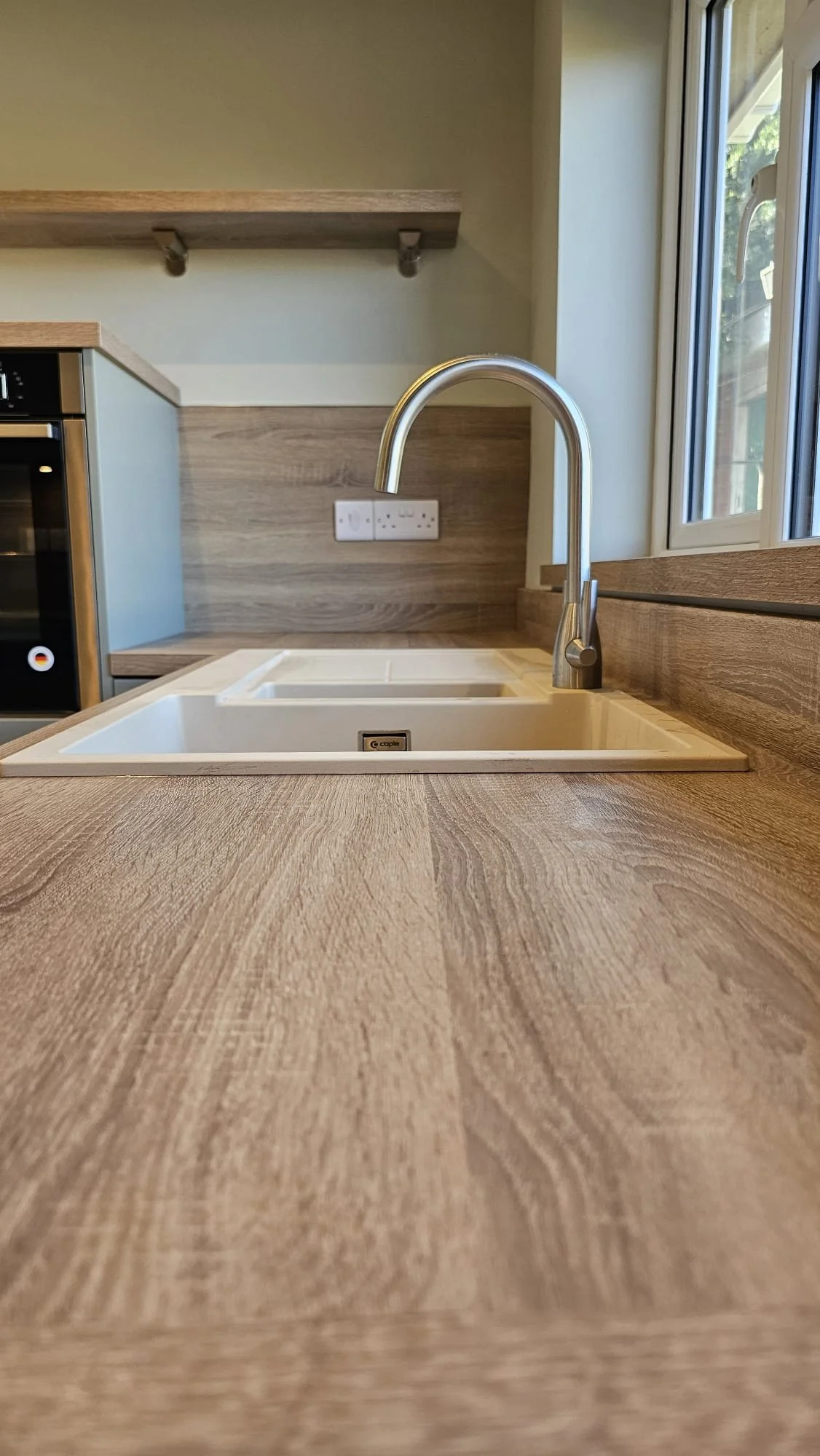 Close-up of a wooden kitchen countertop with a built-in white sink and a modern silver faucet, next to a window.