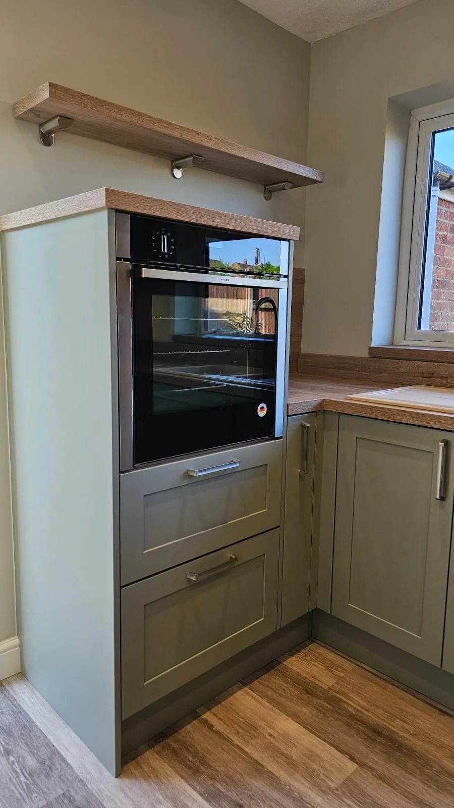 Kitchen with a built-in oven, light green cabinets, wooden countertops, two wooden shelves, and a window showing an outdoor brick wall.