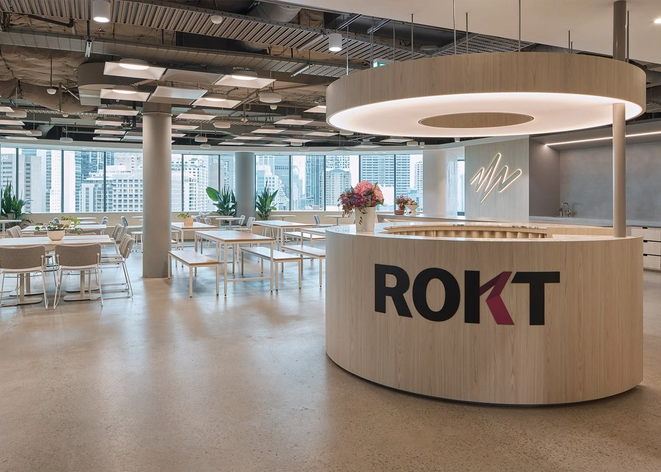Modern office cafeteria with a curved reception desk and large windows showing a cityscape. The space has white tables, chairs, and potted plants, with a light wood finish on the furniture and reception area. Bespoke stretch ceiling lighting custom