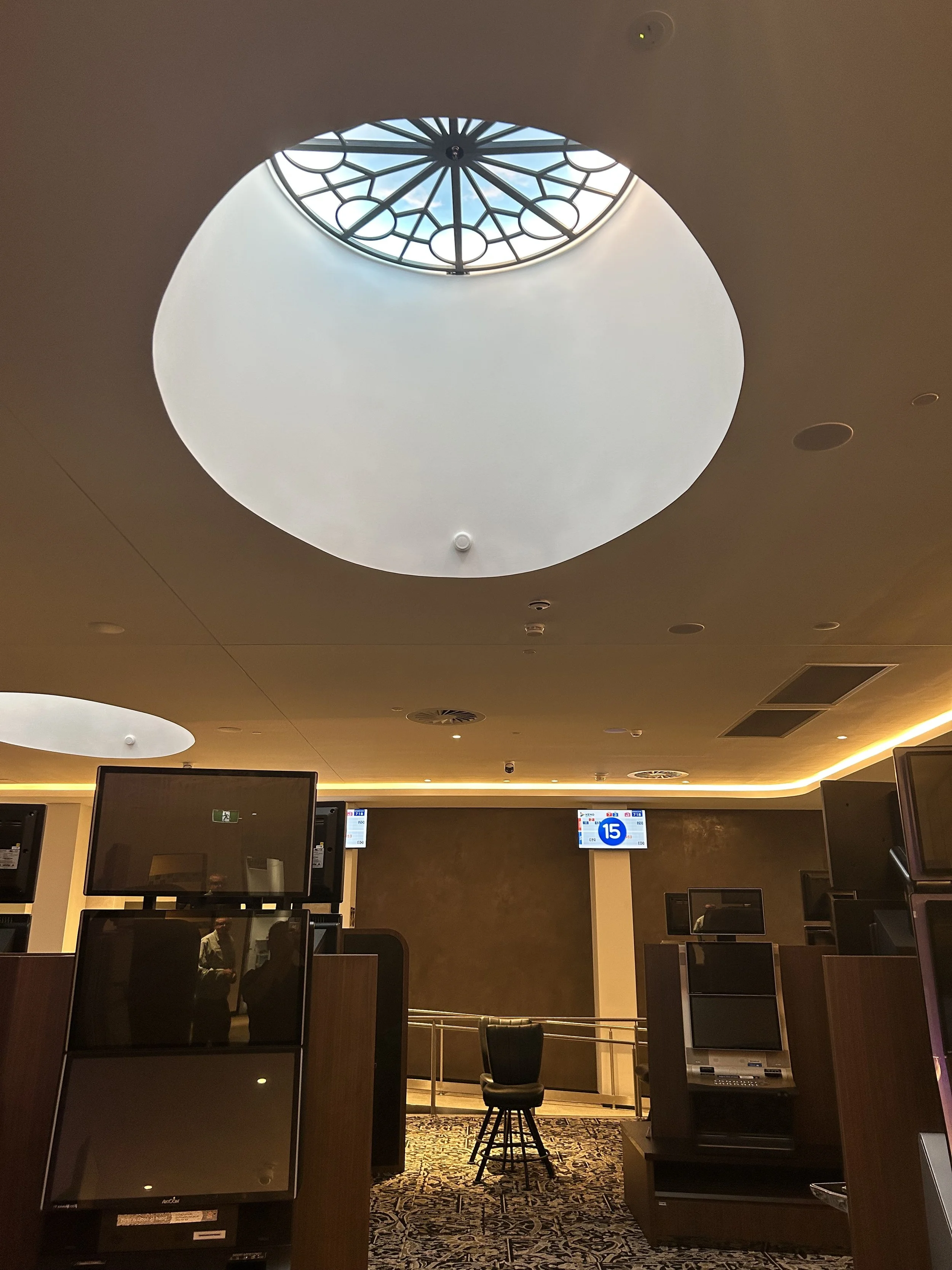 Inside a waiting area with multiple monitor screens, a decorative ceiling with a circular skylight, and a single chair in the center. The area is softly lit with warm lighting. Bespoke stretch ceiling lighting custom designs.