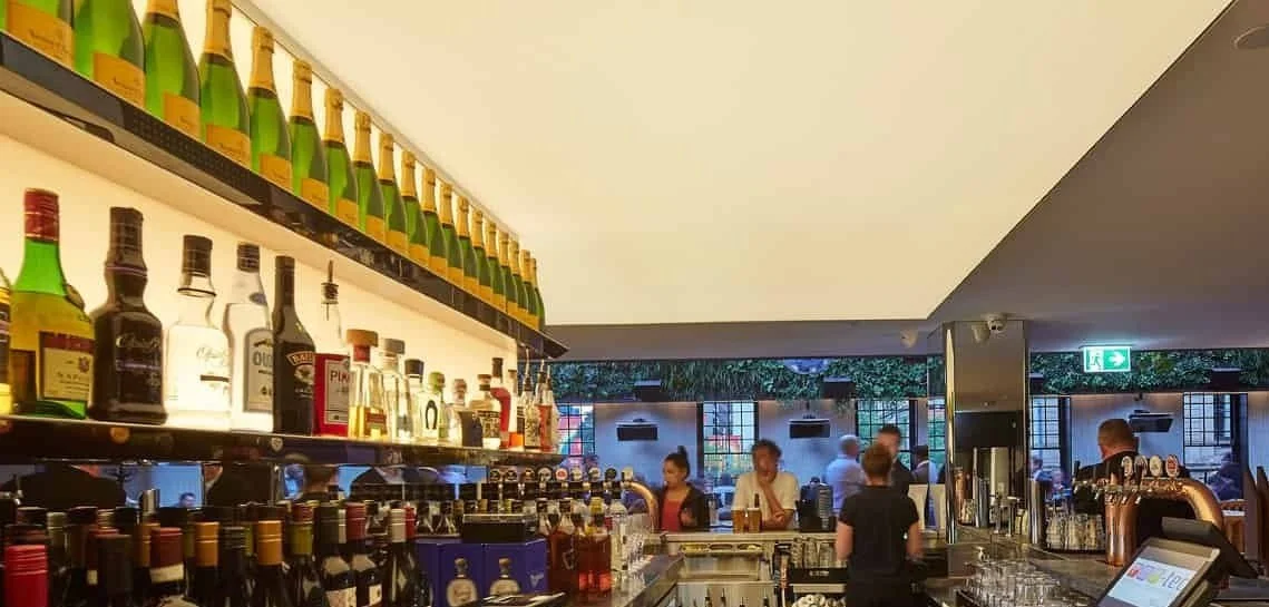 Bar with bottles of alcohol on shelves and bar staff serving customers, in a well-lit restaurant or bar setting. Bespoke stretch ceiling lighting custom designs.
