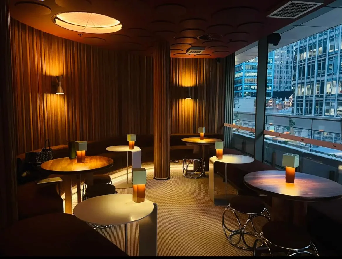 Interior of a modern lounge or restaurant with wooden wall panels, dim lighting, and round tables with small, modern lamps. Large glass windows reveal a city skyline with tall buildings. Bespoke stretch ceiling lighting custom designs.