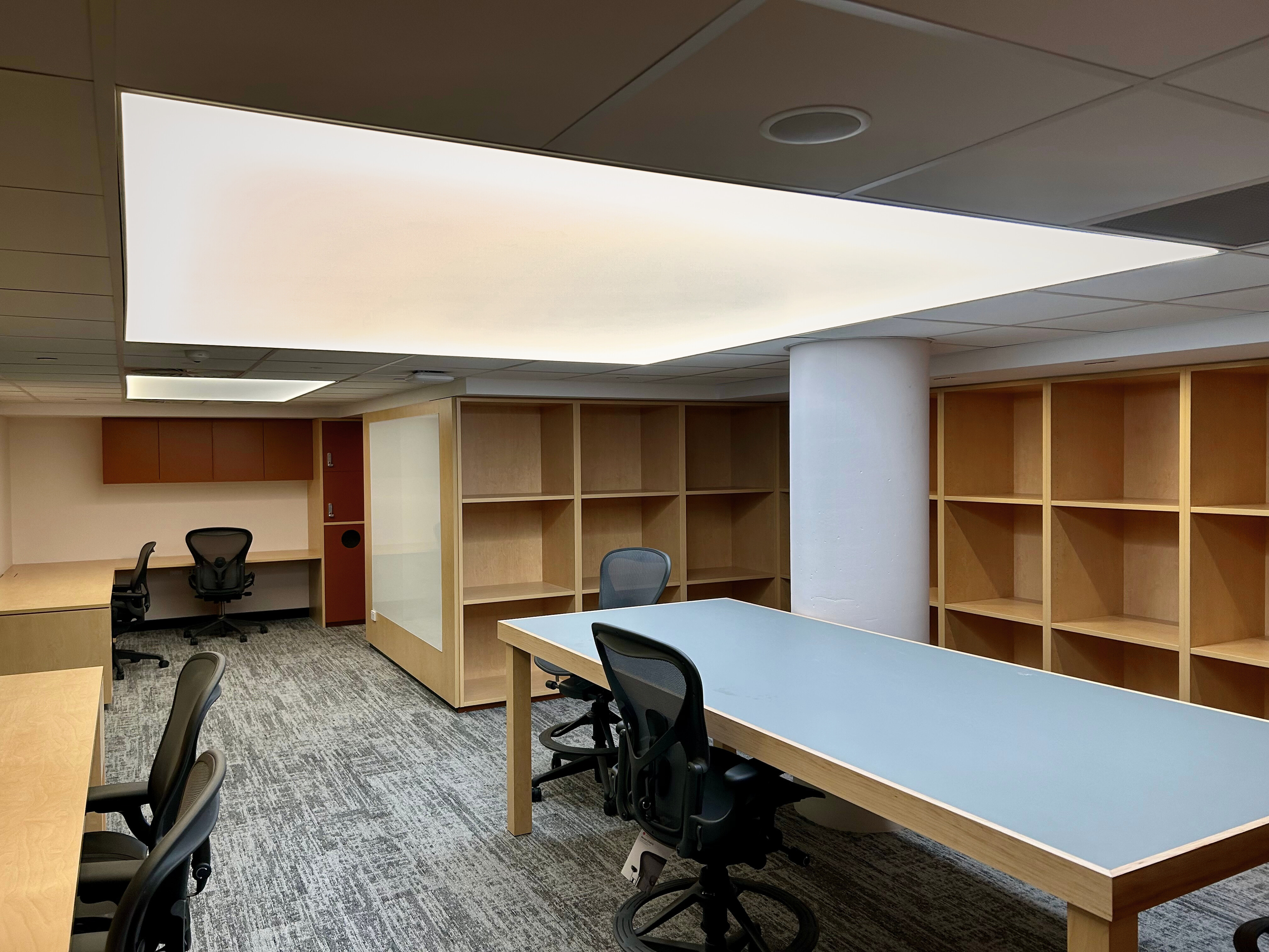 Empty office space with wooden desks, black office chairs, and empty bookshelves, under a large rectangular ceiling light. Bespoke stretch ceiling lighting custom designs.