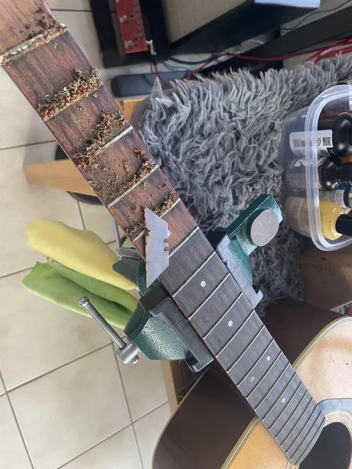 A guitar neck clamped in a work station with a saw blade attached for cutting or shaping, with dirt and debris on the fretboard.