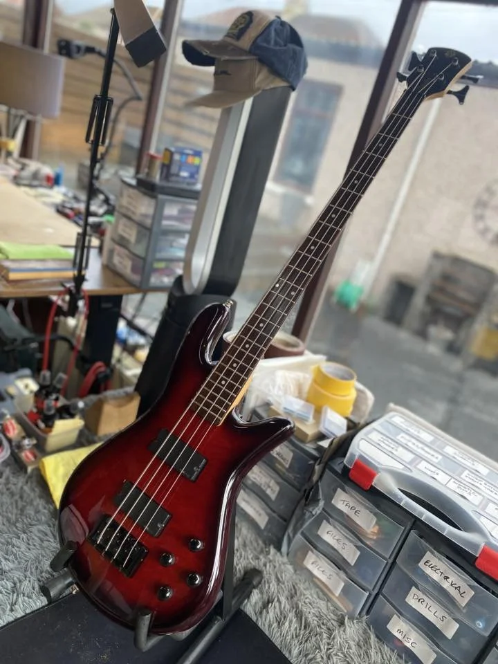 A red and black electric bass guitar with a glossy finish, resting on a stand in a workshop or hobby room surrounded by various tools, parts, and storage containers.