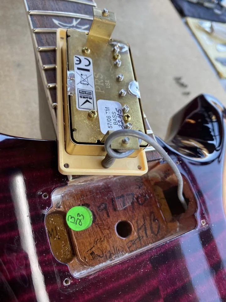 Close-up of the back of an electric guitar body showing the pickup cavity, wiring, and a metal pickup installed, with handwritten notes and a green sticker on the surface.
