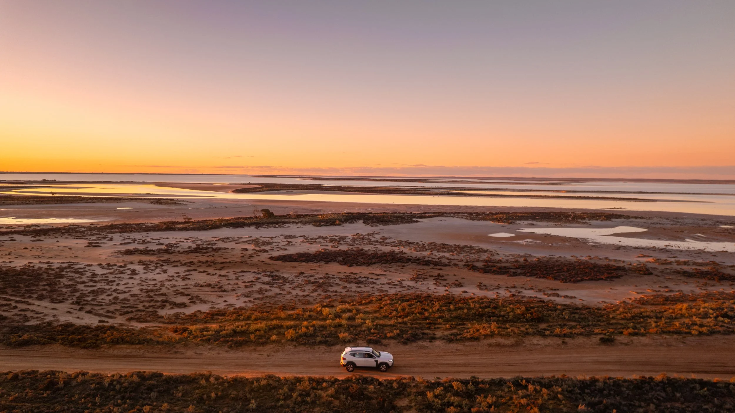 Aerial view of a car driving beside Lake Tyrrell at sunset, with shallow saltwater and golden light across the Mallee landscape in Victoria.