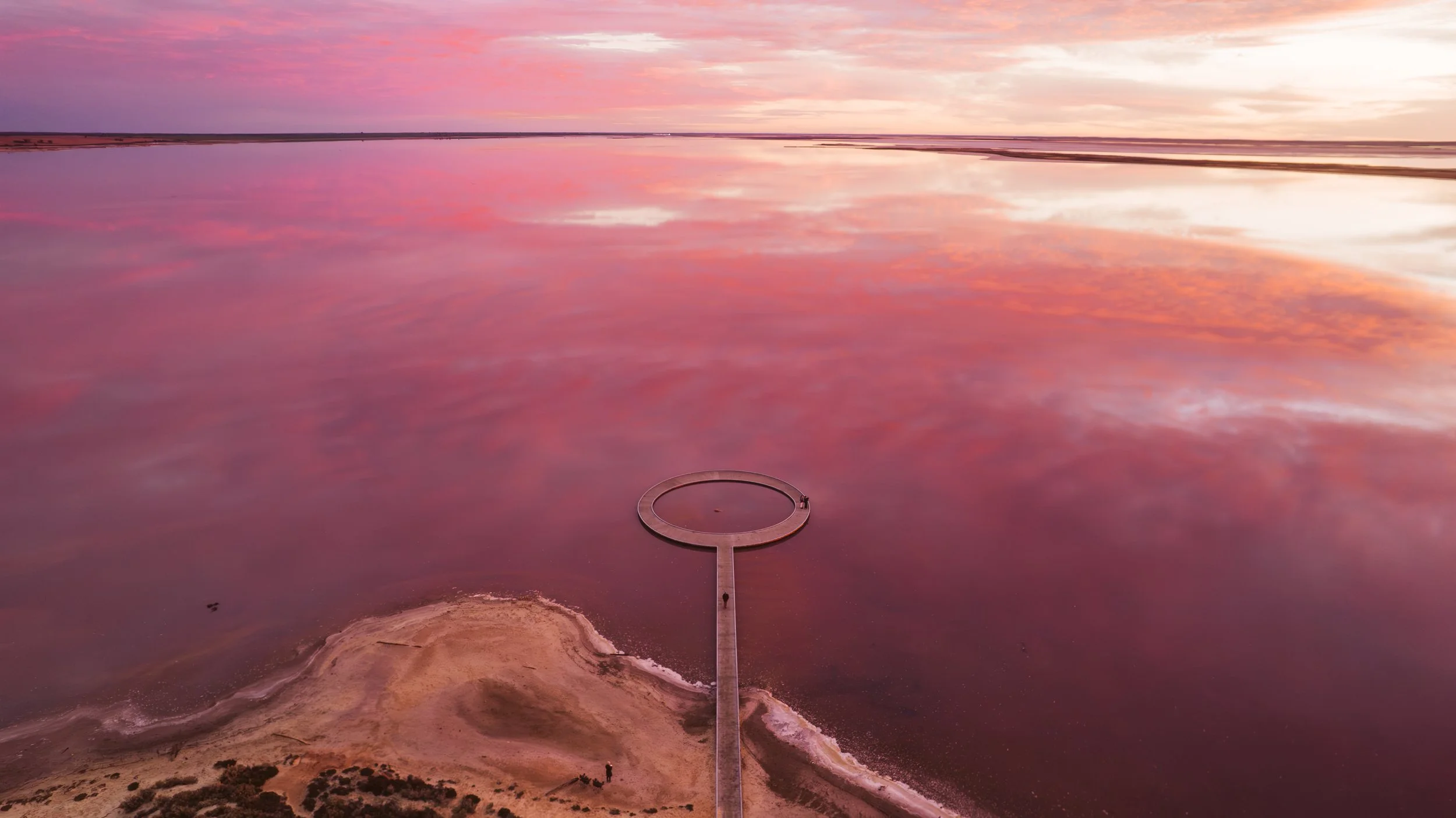 Aerial view of Lake Tyrrell in Victoria at sunrise, with pink salt water reflecting the sky and a circular viewing platform extending into the lake.