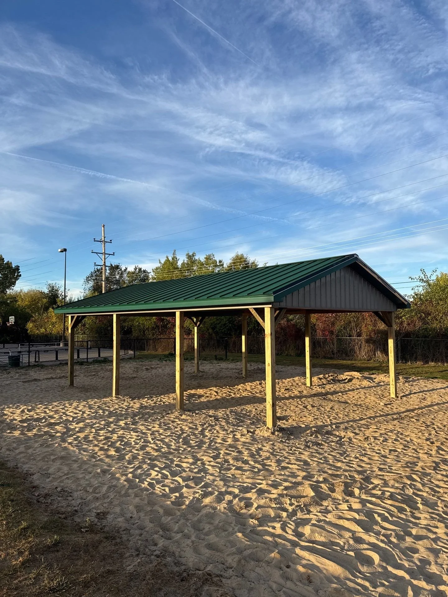 A small pavilion with a green metal roof on a sandy area, with trees and power lines in the background, under a partly cloudy sky.