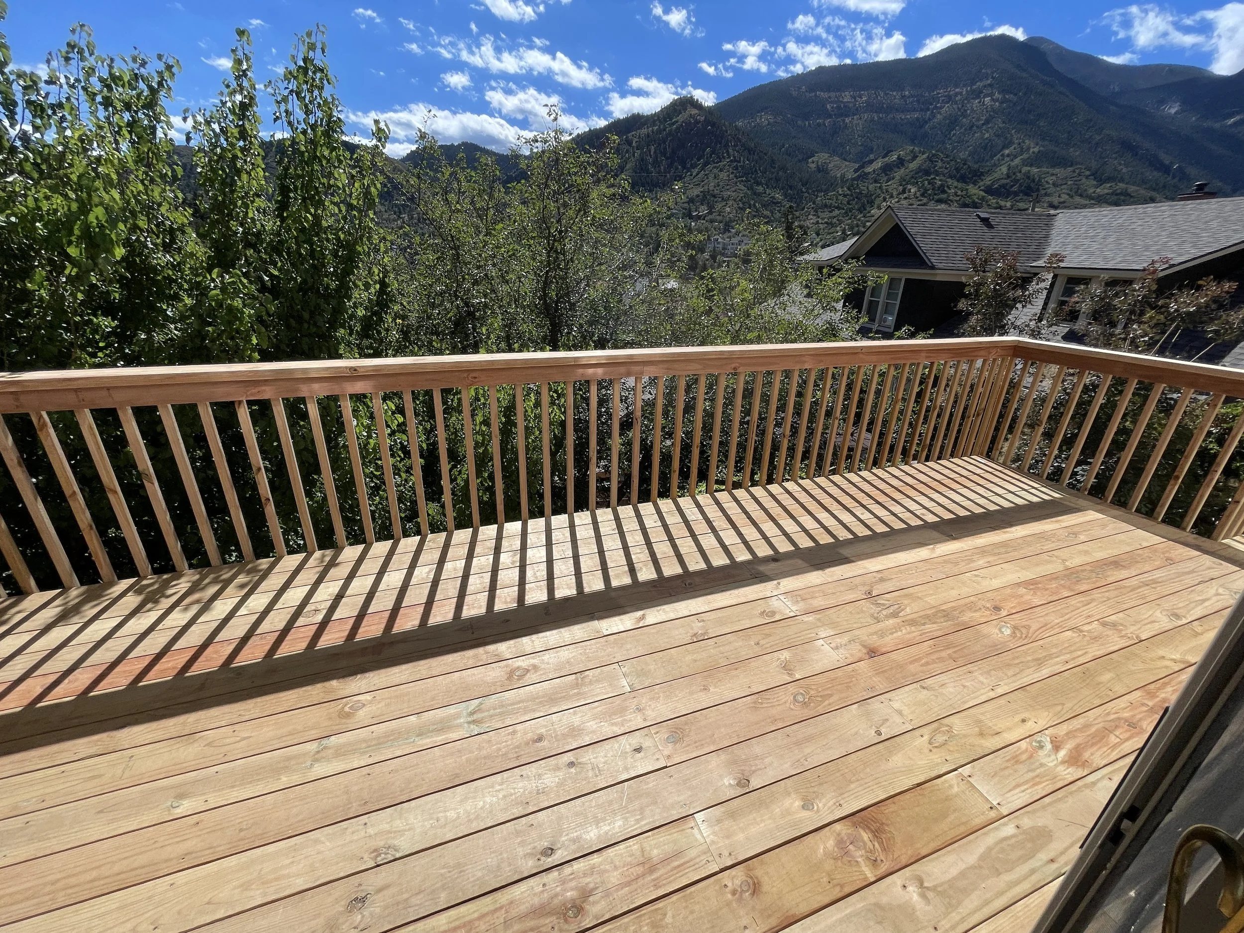 Wooden deck with railing overlooking trees and mountains under a partly cloudy sky.