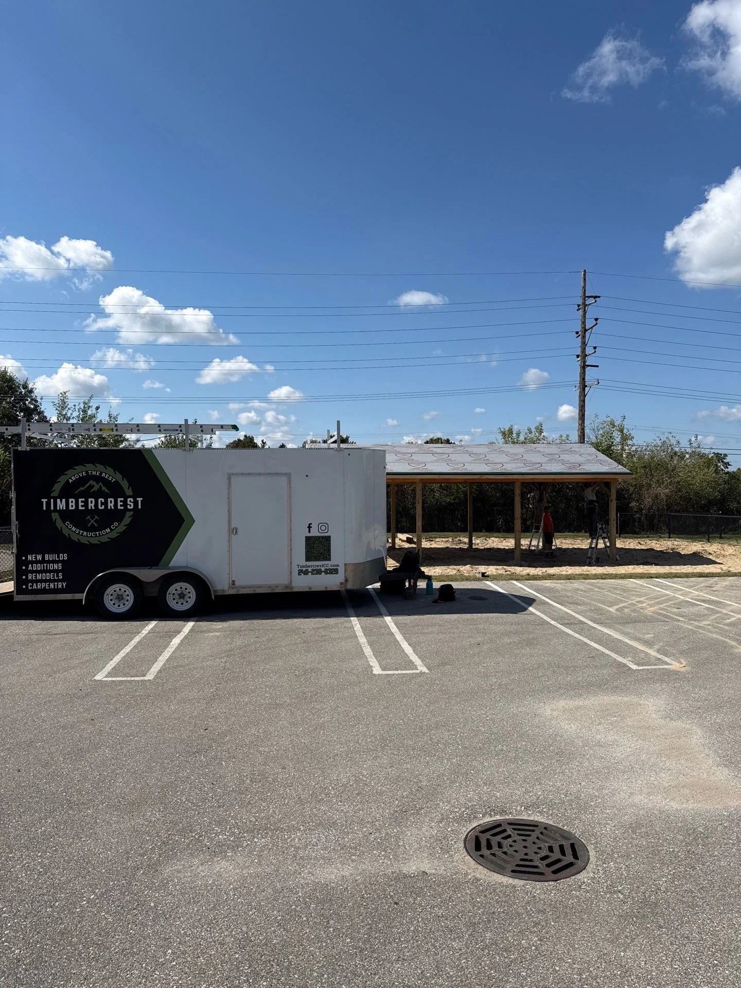 Construction site with a Timbercrest Construction trailer and a small open frame building under construction, with workers and construction tools visible.