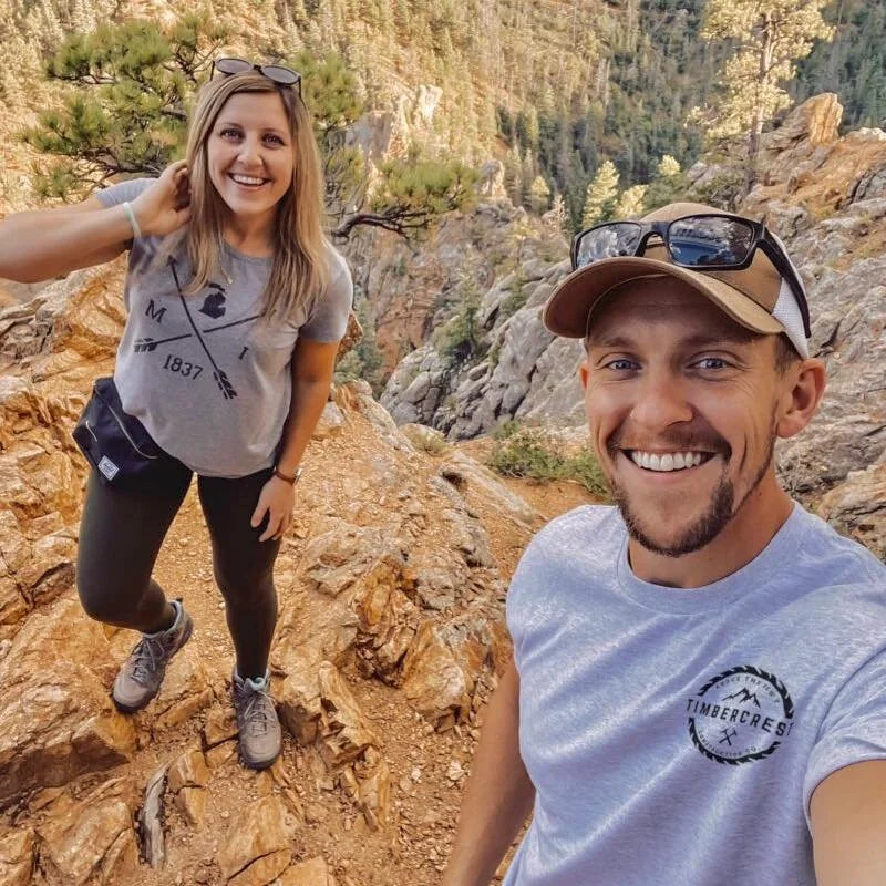 A smiling young woman and man hiking on a rocky trail in a mountainous, forested landscape during daytime.