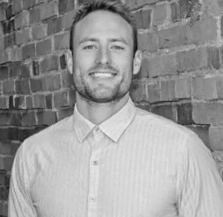 A man smiling in front of a brick wall, wearing a light-colored collared shirt.