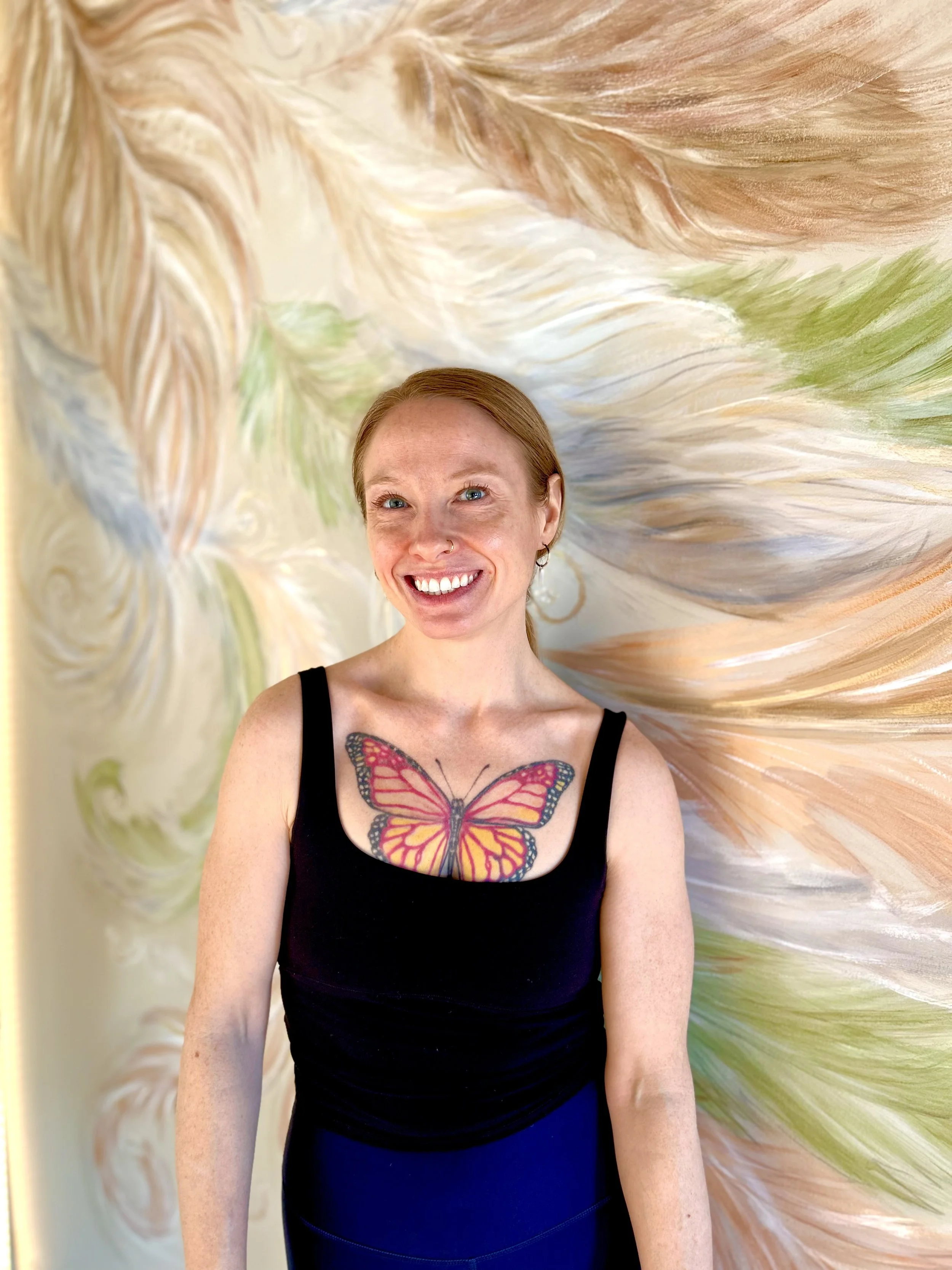 Close-up of a woman with red hair, glasses, and a cheerful smile, outdoors with a blurred green background.