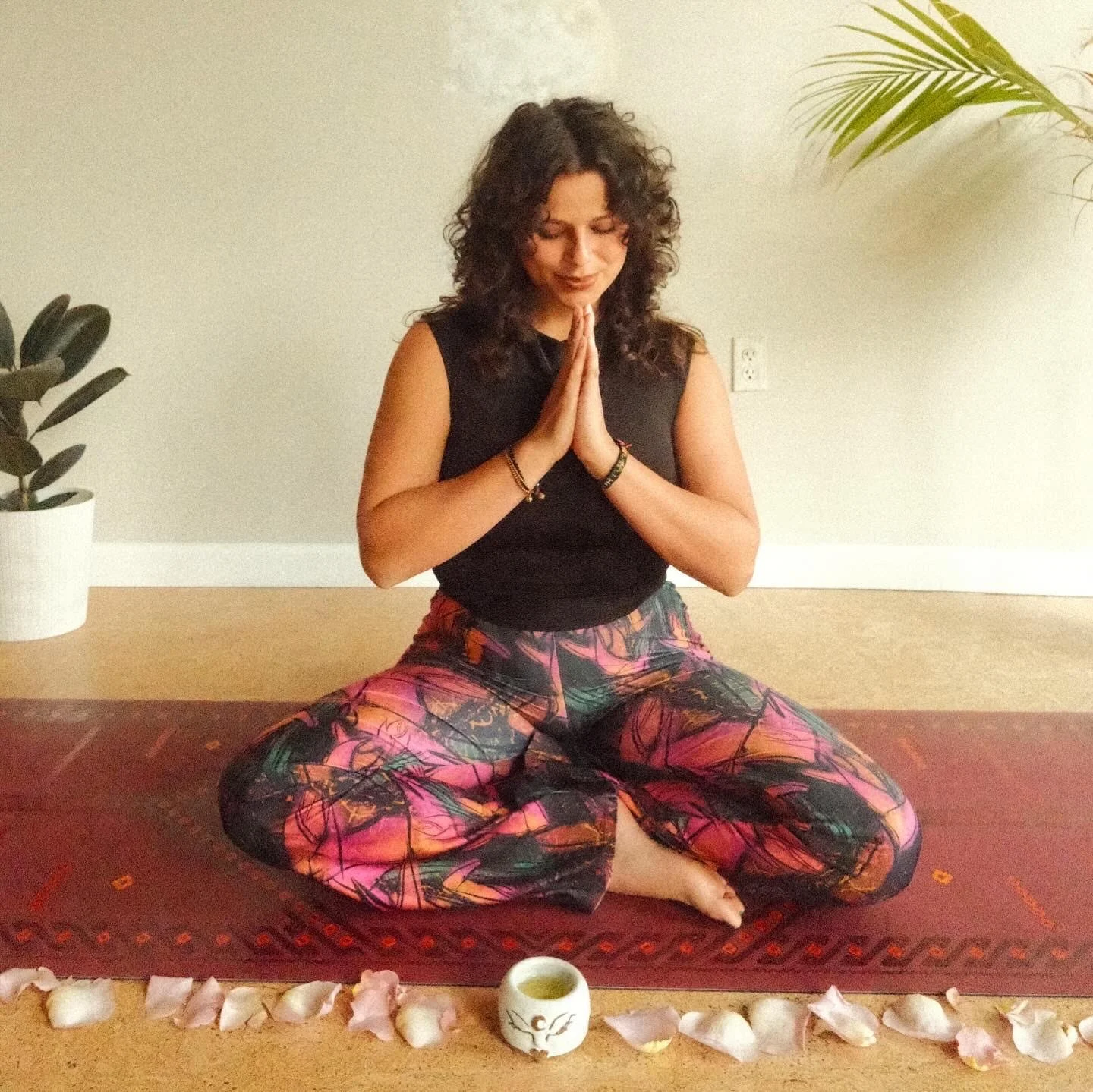 A woman with shoulder-length curly brown hair, wearing a sleeveless brown top and light-colored pants, leaning on a wooden surface in a warmly lit indoor setting.