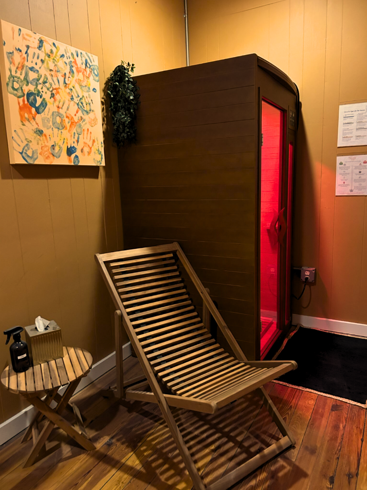 A wooden room features a wooden lounge chair, a small wooden side table with a bottle of disinfectant spray and tissues, a colorful abstract painting, a hanging green plant, and a large wooden sauna with red interior lighting.